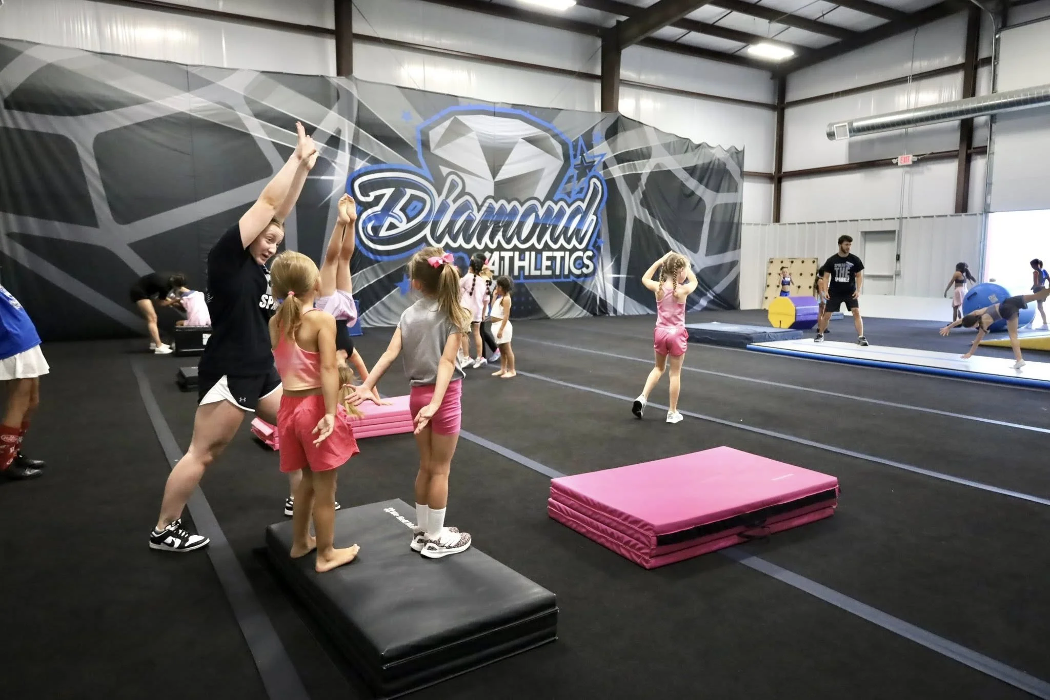 Kids practicing gymnastics in an indoor gym, with a large wall display reading 'Diamond Athletics' in the background, and various gymnastic equipment like mats, foam blocks, and balance beams.