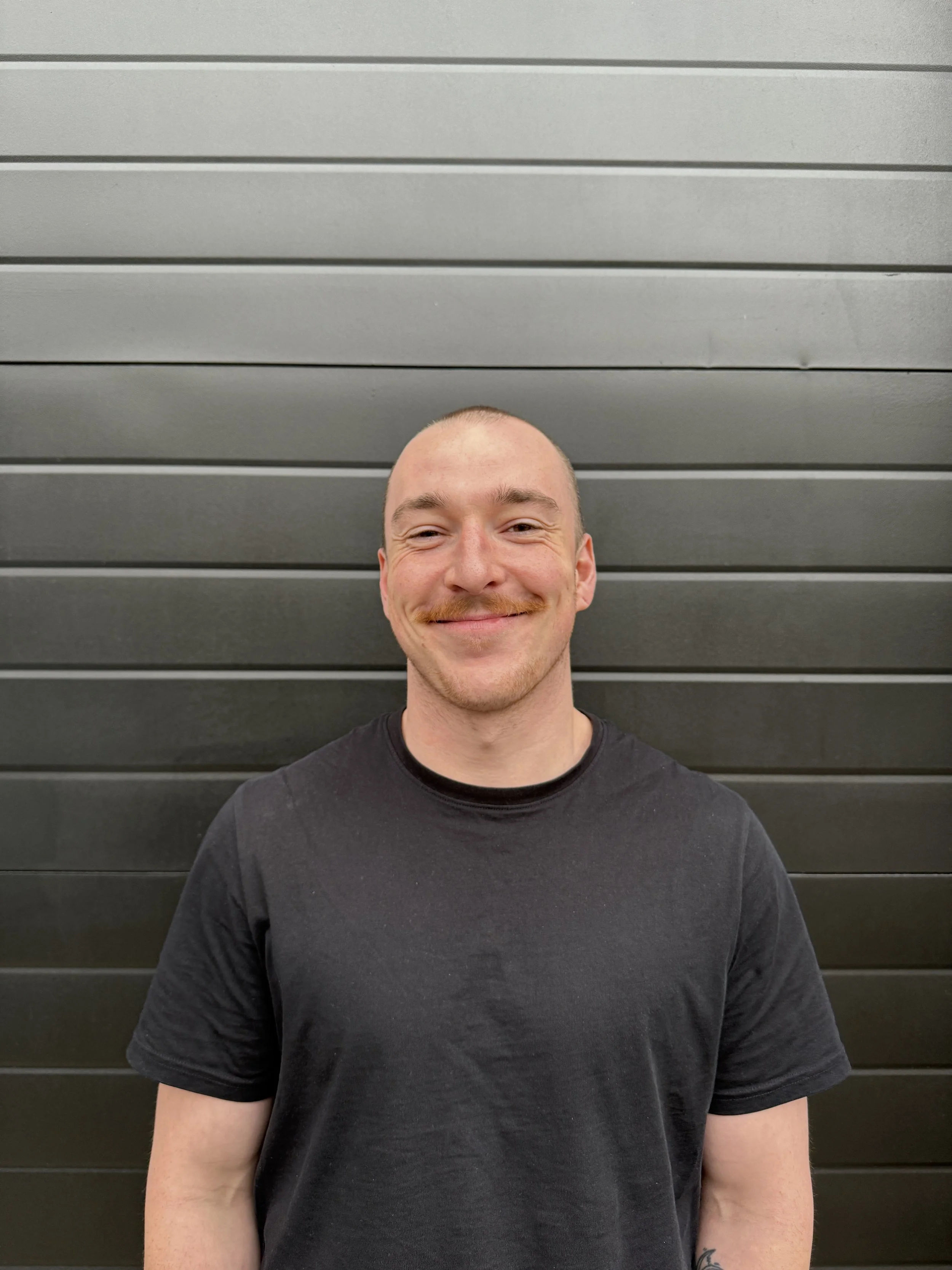 A man with a mustache, smiling, standing in front of a black horizontal panel wall. He is wearing a black T-shirt.