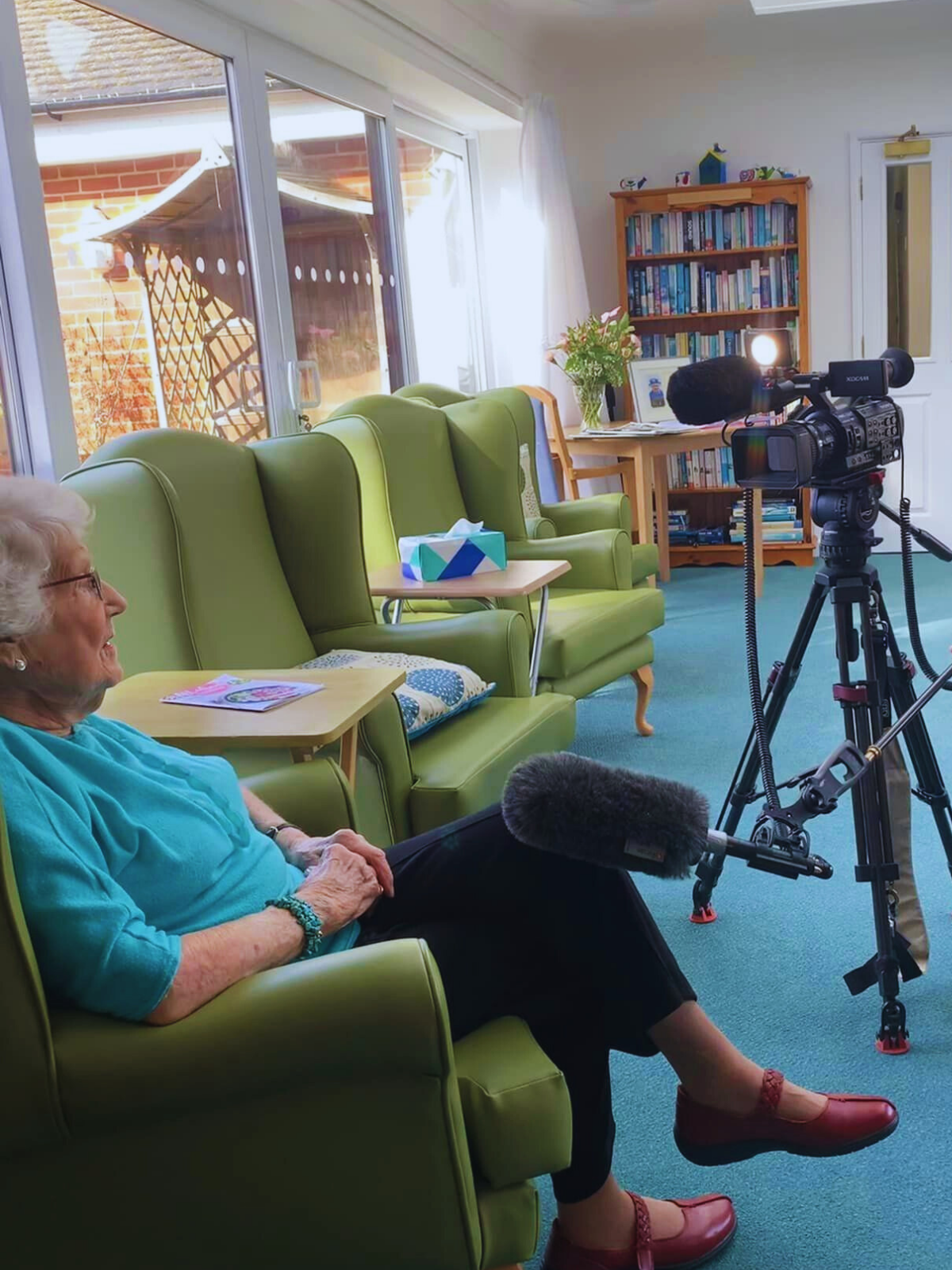 An elderly woman sitting on a green armchair inside a well-lit room, with a camera set up on a tripod in front of her. The room has large windows, a bookshelf filled with books, a table with a bouquet of flowers, and green armchairs.