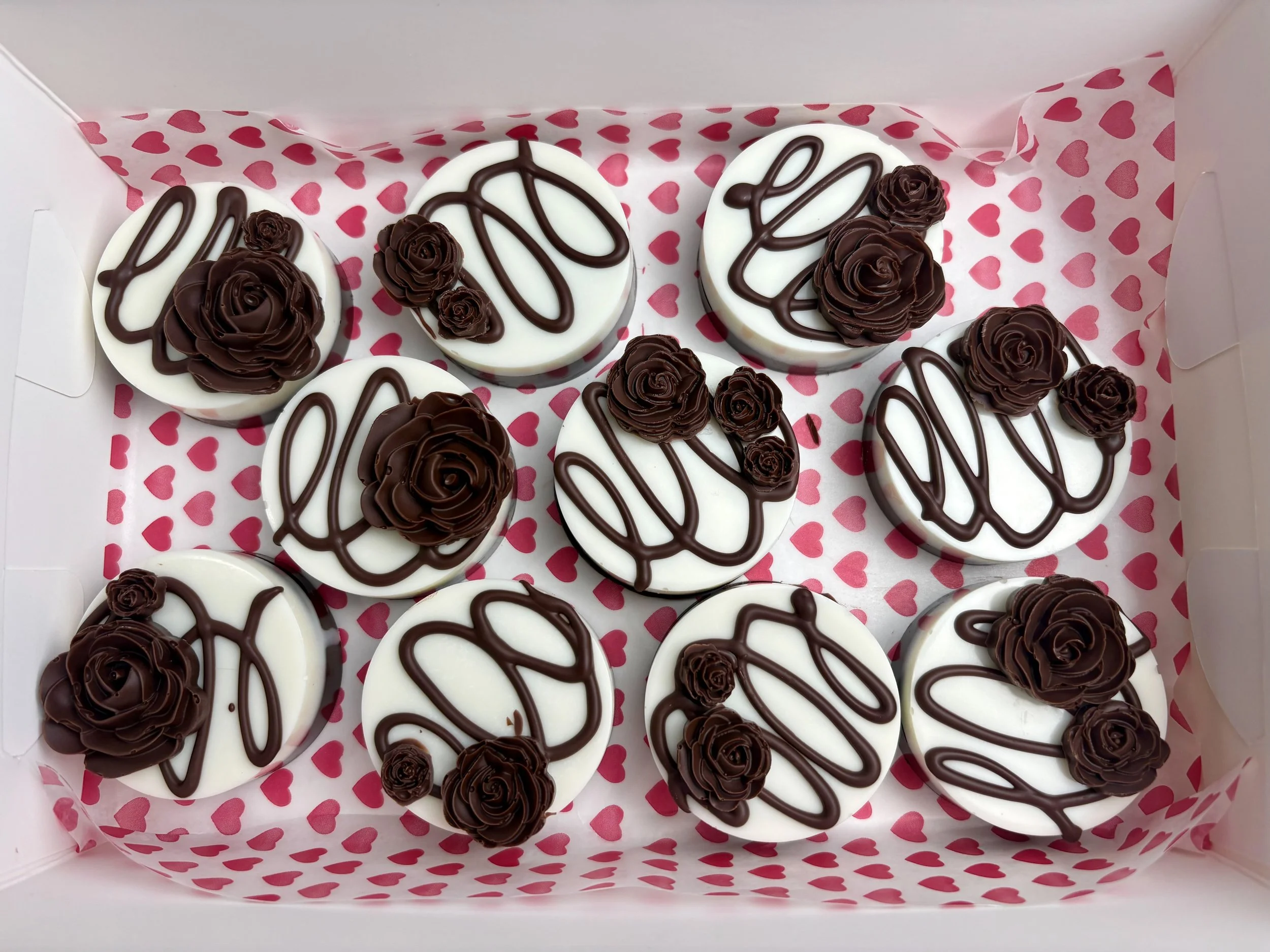 Box of decorated cupcakes with white frosting, drizzled with chocolate, topped with chocolate roses, inside a white box with pink heart-patterned paper lining.