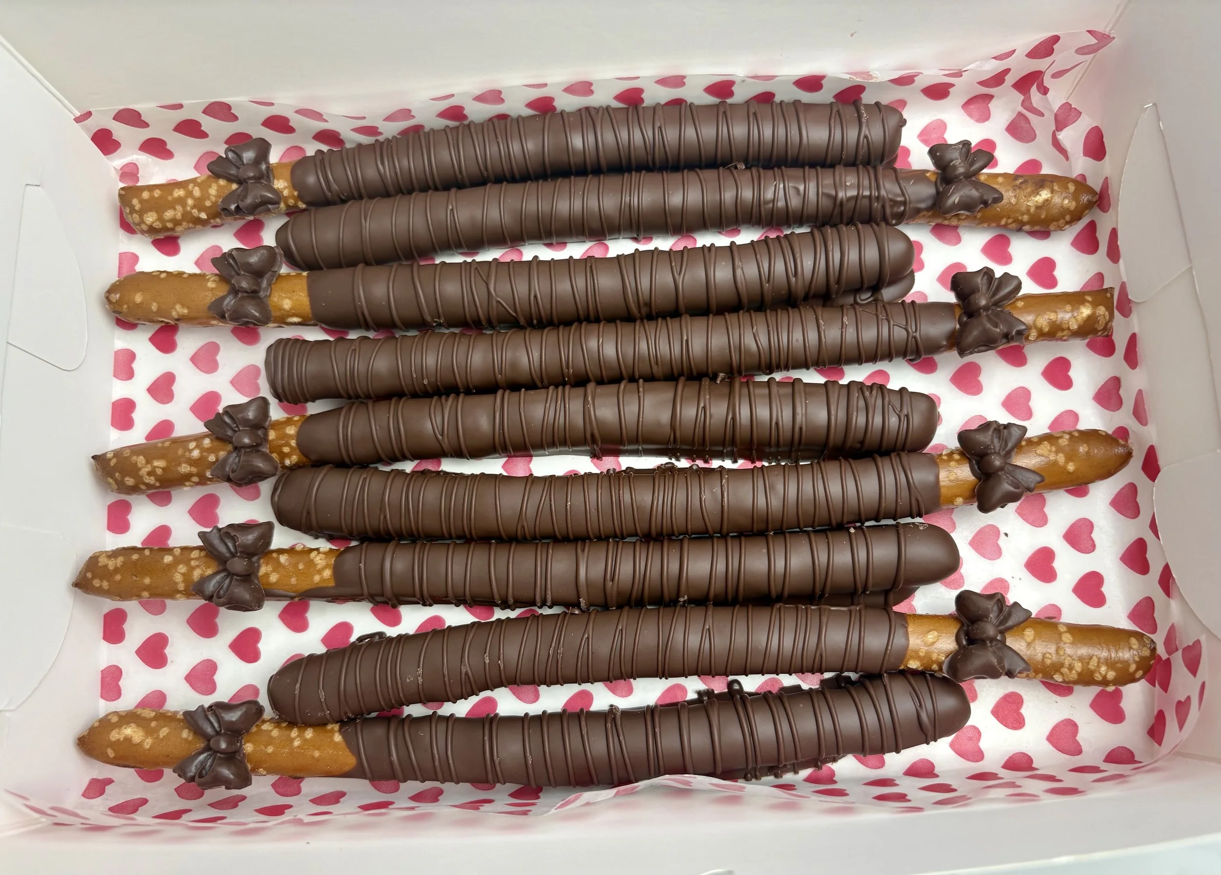 A box of chocolate-covered pretzel rods decorated with small chocolate bows on the ends, arranged on pink heart-patterned paper.