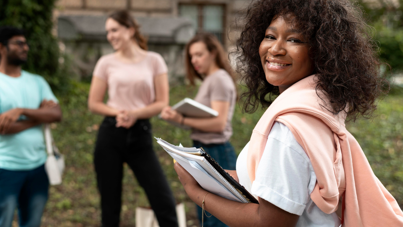 Smiling young woman holding notebooks outdoors with a group of four people in the background.