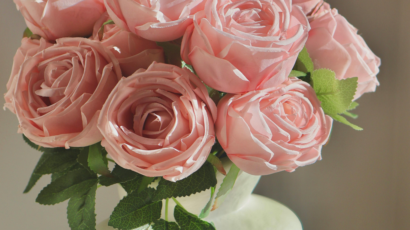 Close-up of a bouquet of pink artificial roses in a white vase, with green leaves surrounding the flowers.