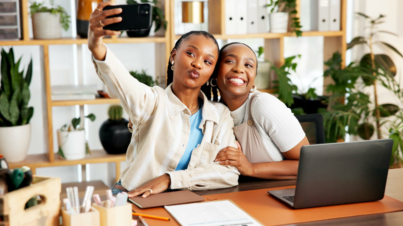 Two women taking a selfie together while sitting at a desk in an office or home workspace, with a laptop and office supplies in front of them, surrounded by green plants and shelves with books and decor.