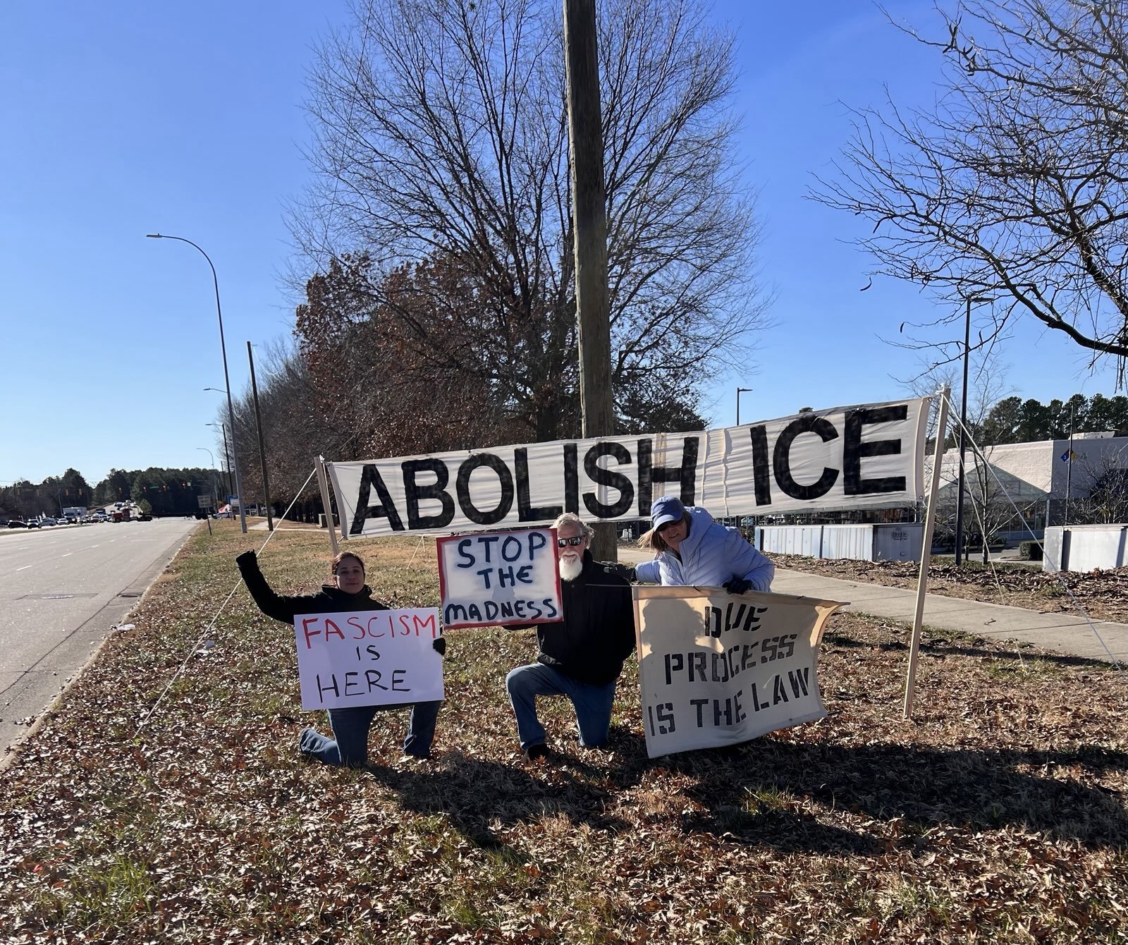 Three people holding protest signs in front of a large banner that reads 'ABOLISH ICE' on a grassy sidewalk next to a street, with leafless trees and a blue sky in the background.