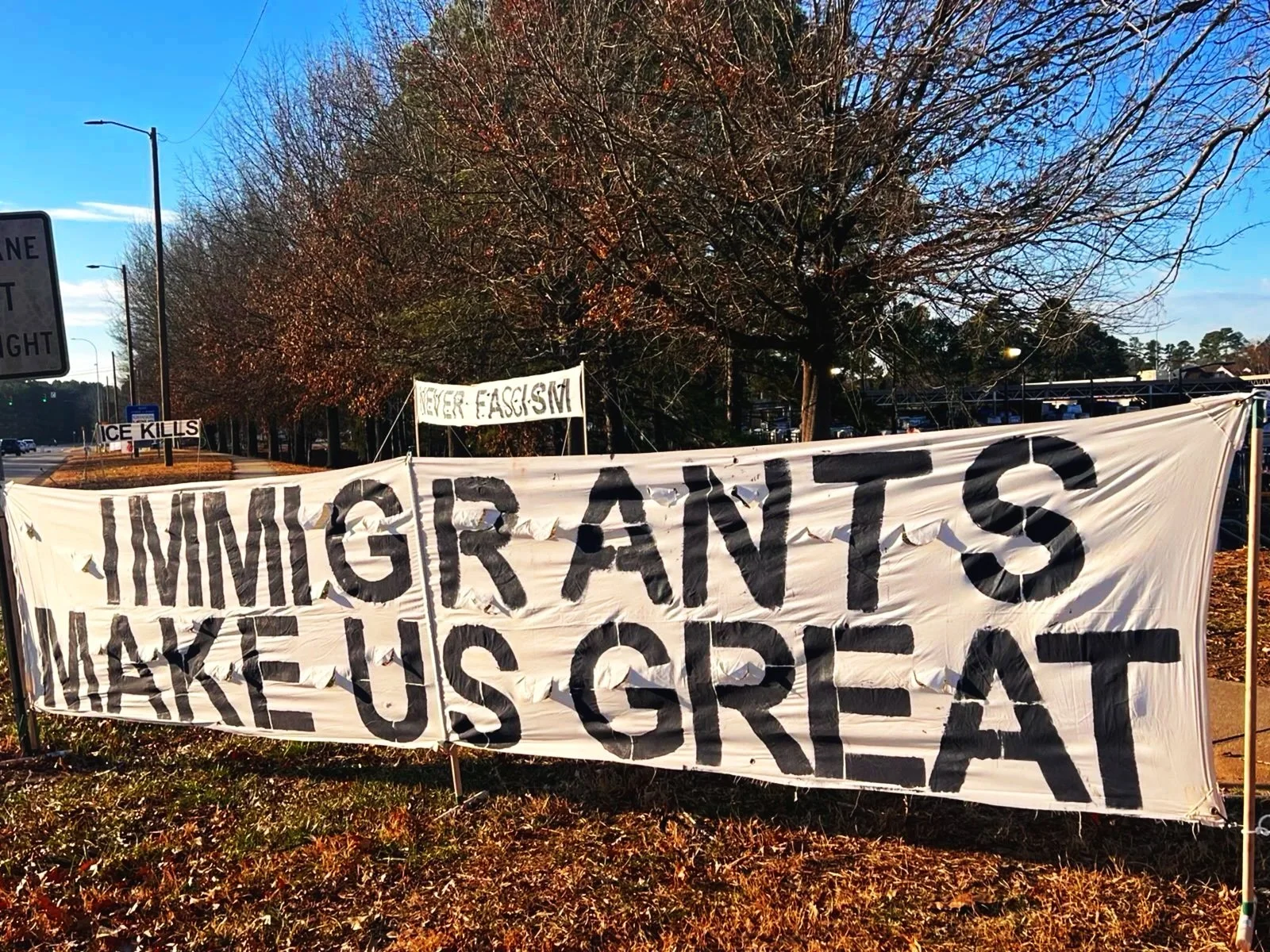 Protest signs on a grassy area by the road, including a large banner reading 'IMMIGRANTS MAKE US GREAT' and smaller signs stating 'ICE KILLS' and 'NEVER FASCISM.'