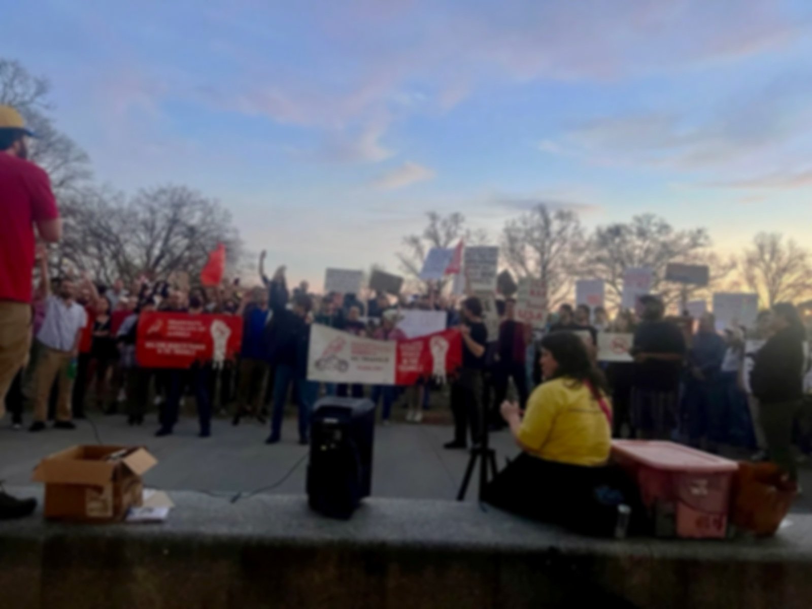A large group of protesters gather outdoors during sunset, holding signs and banners, with some waving fists. There are trees in the background and a woman in a yellow jacket sitting in the foreground near audio equipment.