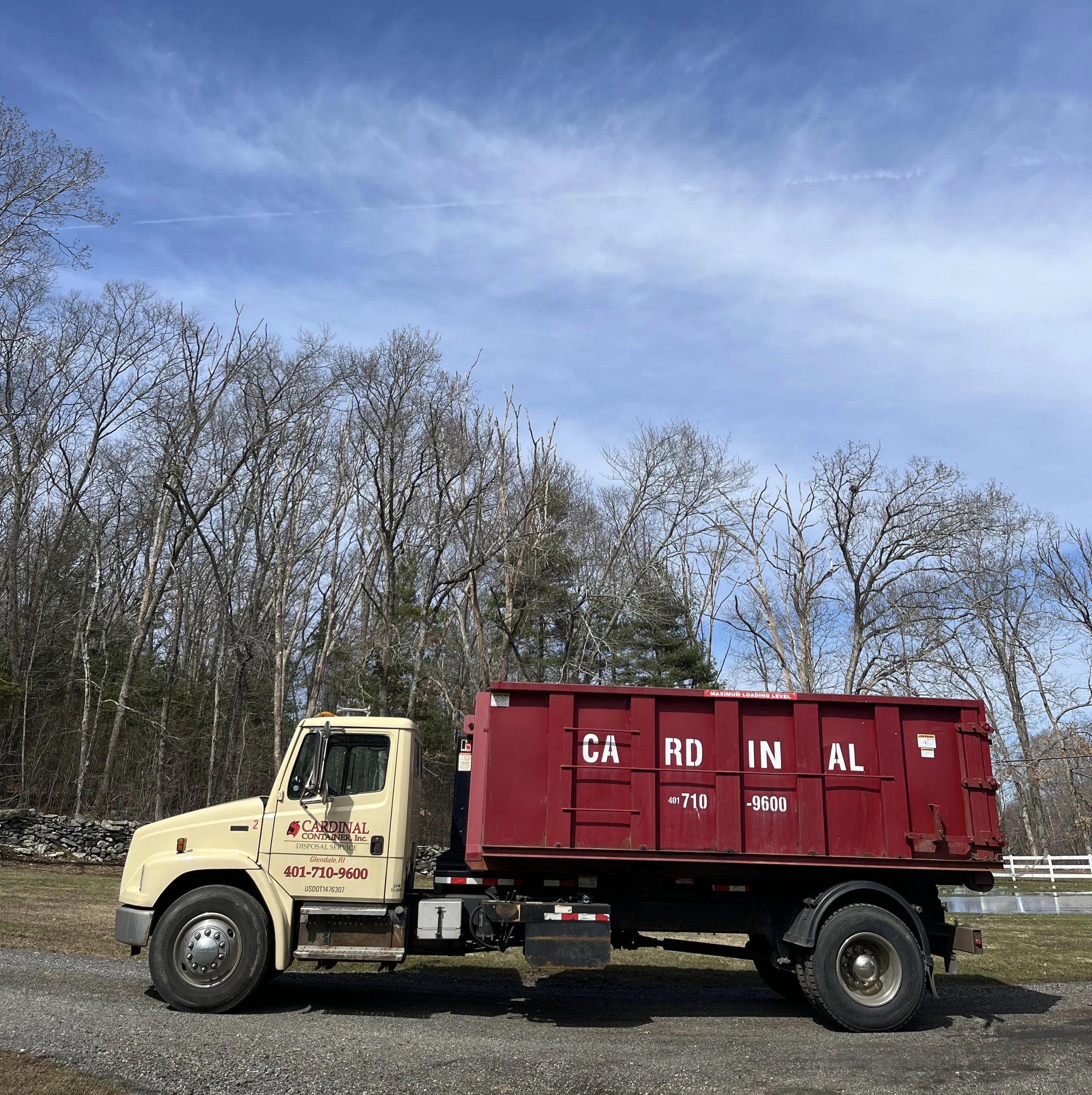 A beige disposal truck with red container in a rural outdoor setting with leafless trees and a blue sky.