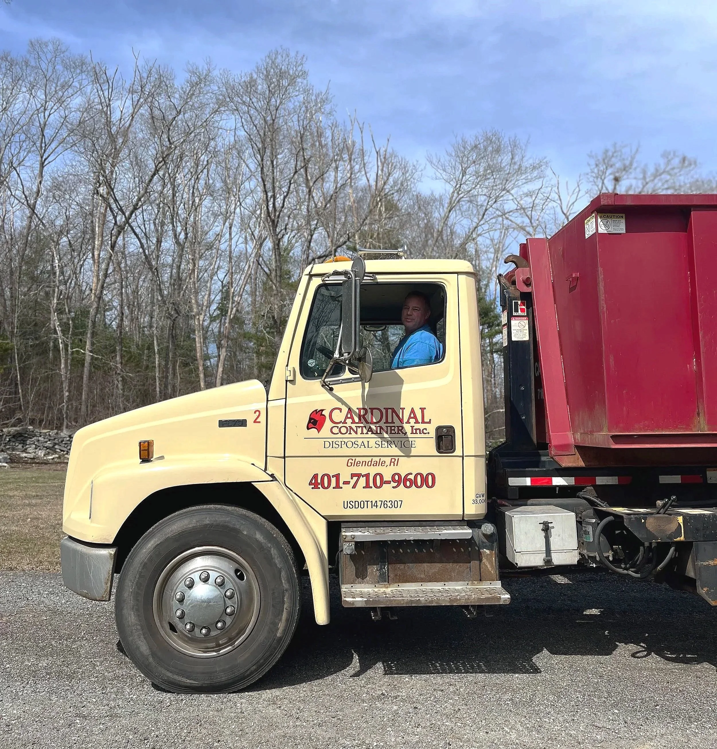 A man sitting in the driver's seat of a light yellow Cardinal Container disposal truck with a red dumpster attached, parked on a gravel surface with leafless trees and a partly cloudy sky in the background.