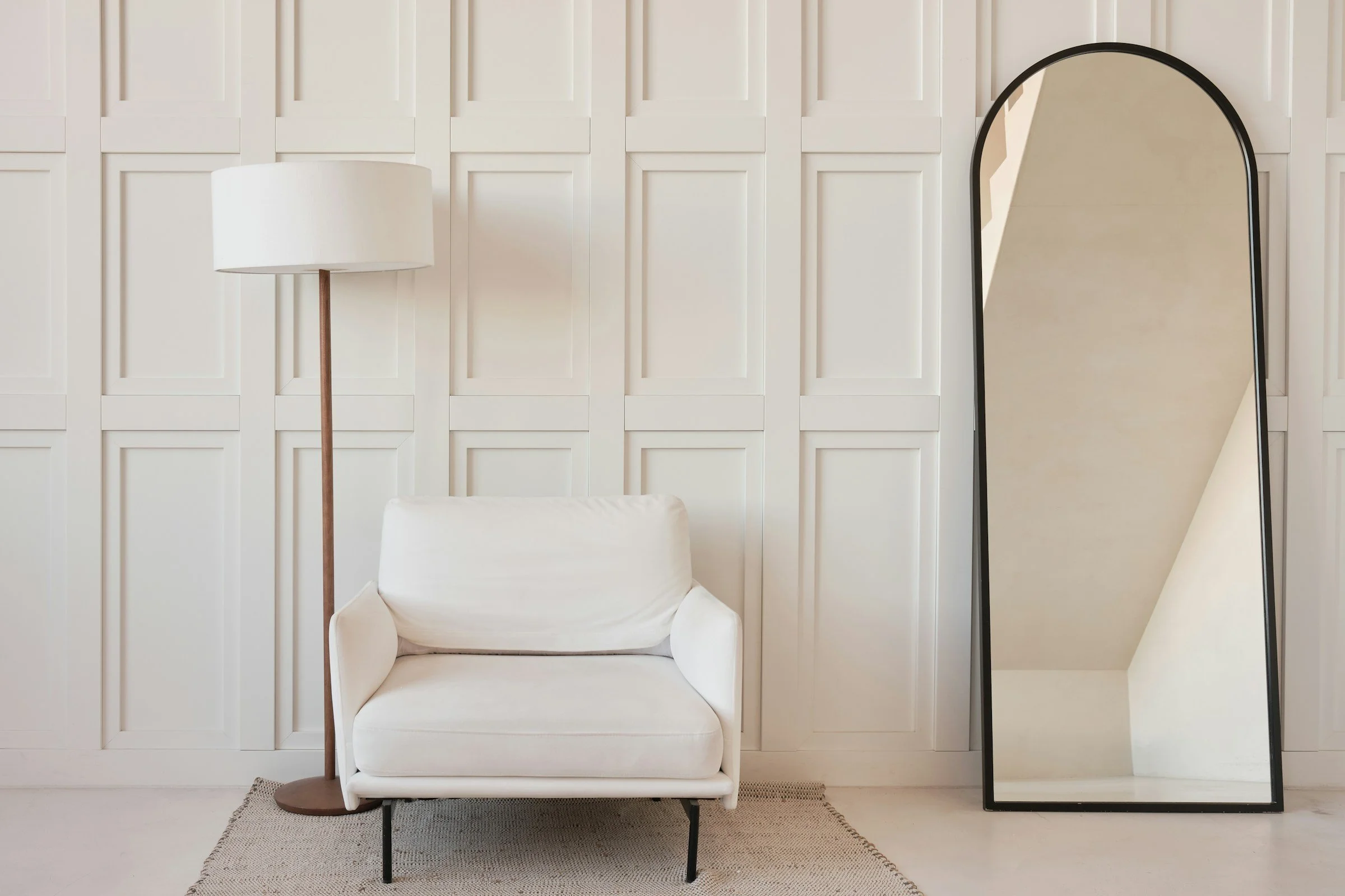 A white armchair, a tall white floor lamp, and a large black-framed mirror leaning against a paneled white wall in a minimalist room.