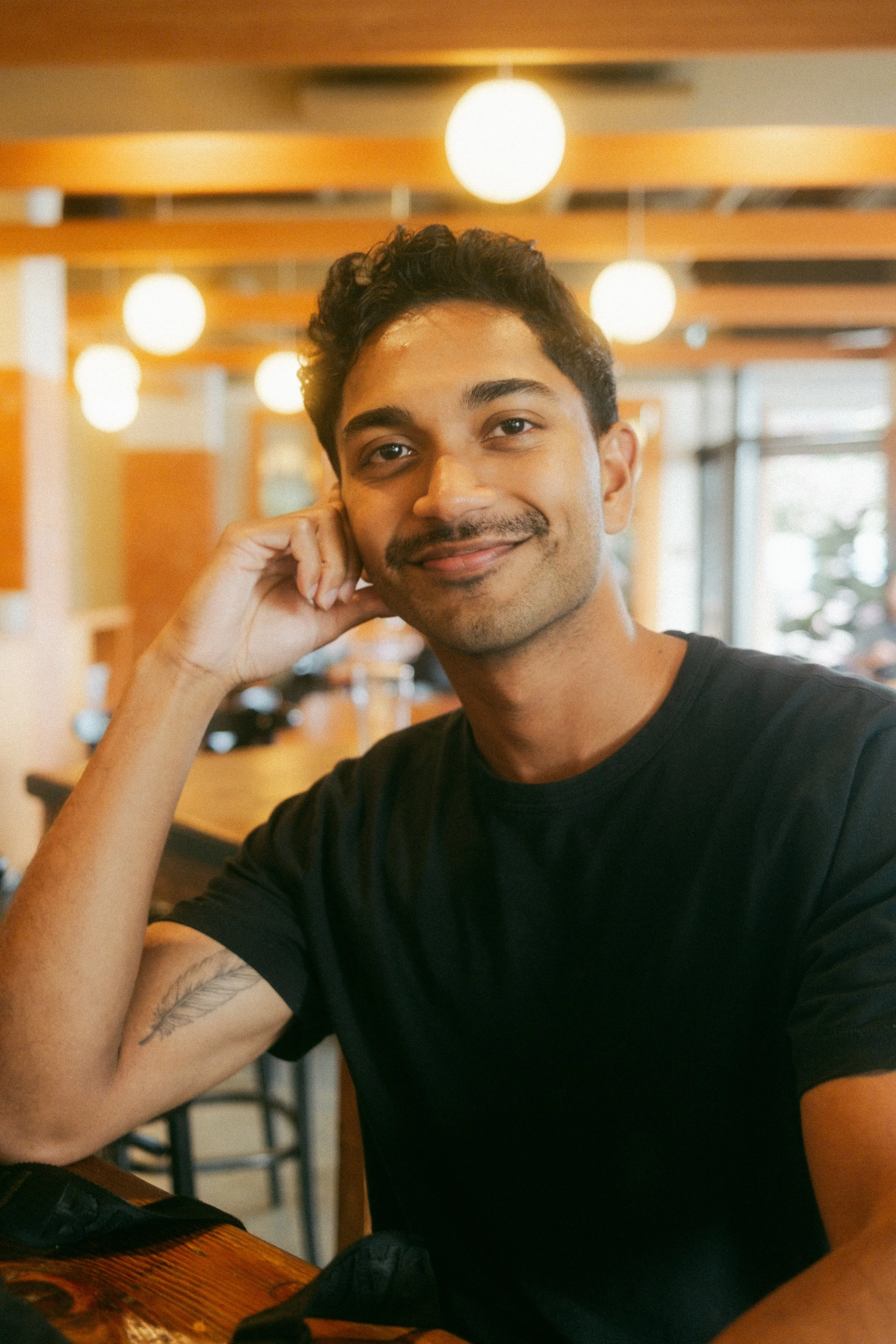 A young man with dark hair and a mustache smiling with his head resting on his hand in a well-lit cafe or restaurant.