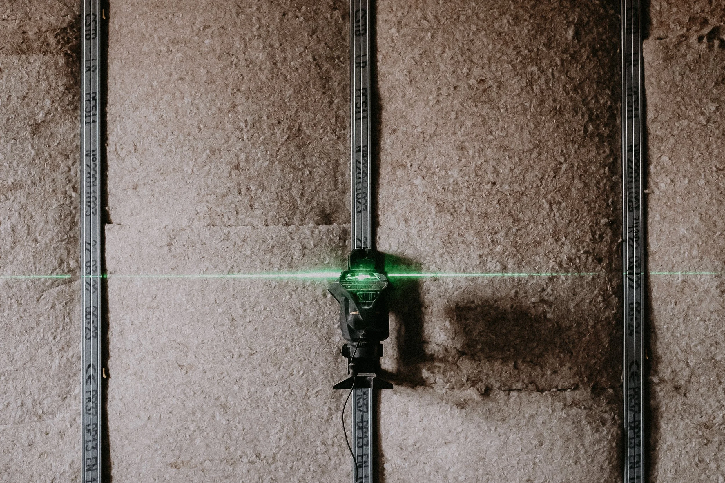 Laser level mounted on a wall with exposed drywall panels and metal tension strips.