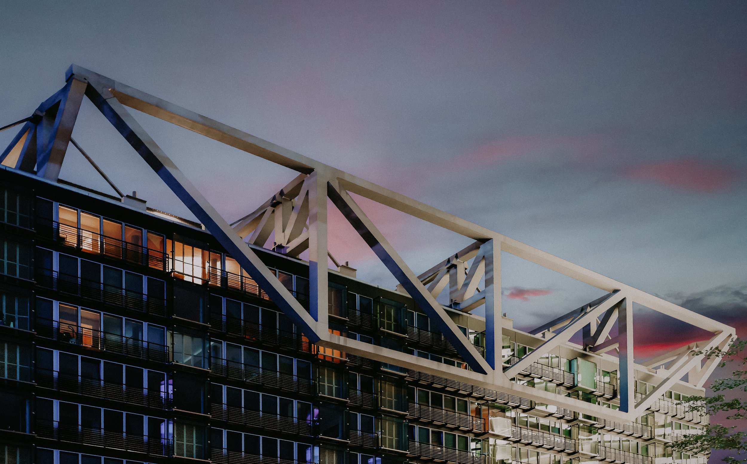Modern multi-story building with geometric architectural features on the roof, illuminated windows, and a dusk sky with pink and purple clouds.