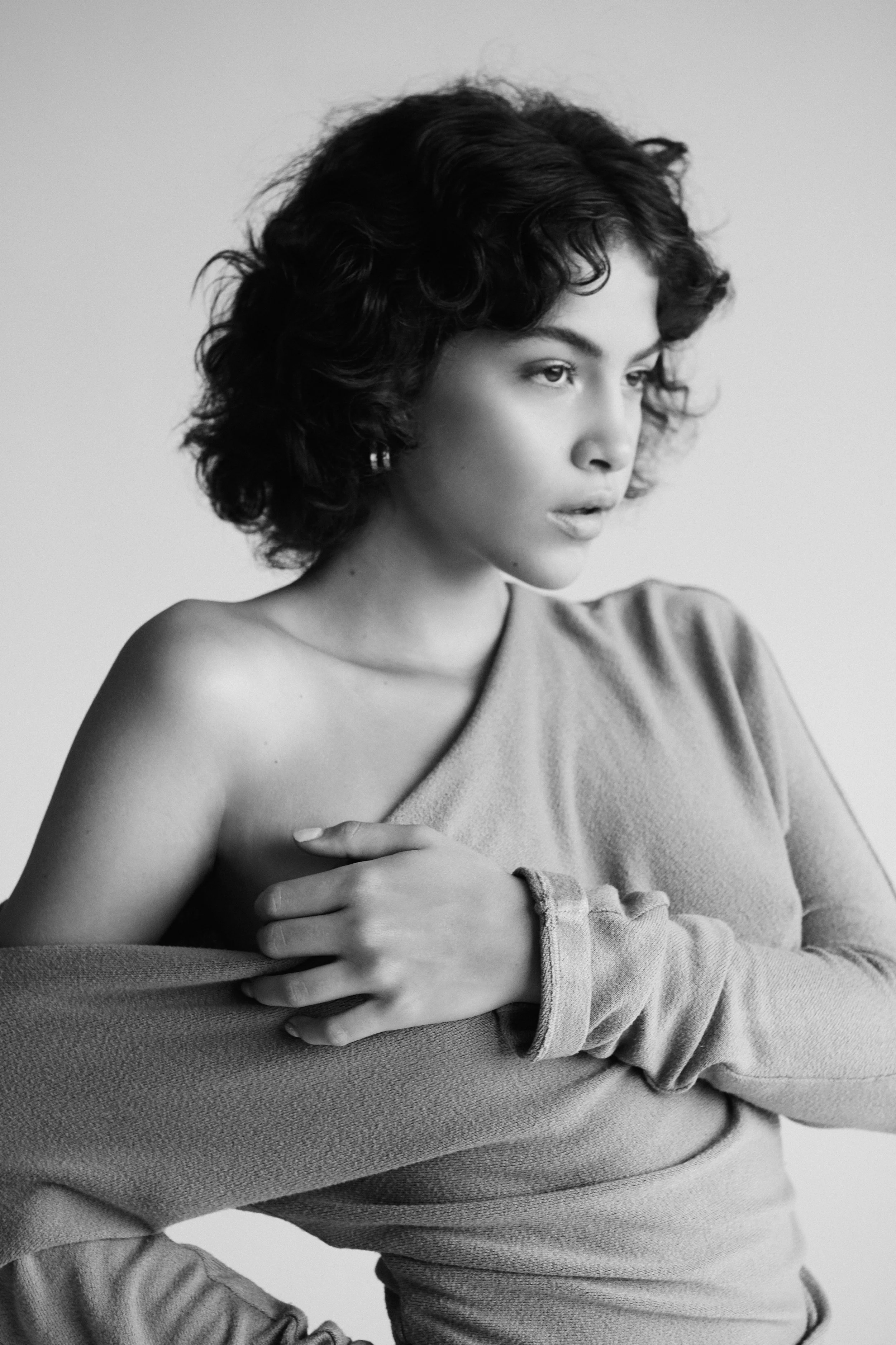 Black and white portrait of a young woman with curly hair wearing a one-shoulder top, looking to the side.