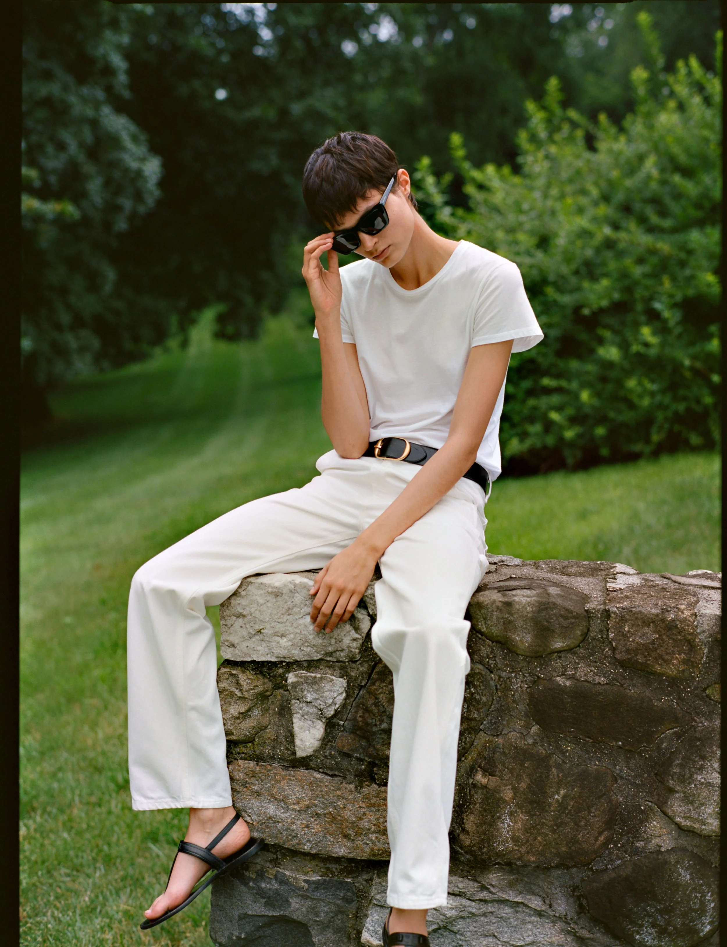 A person wearing black sunglasses, white t-shirt, white pants, and black sandals, sitting on a stone wall in a park with green trees and grass in the background.