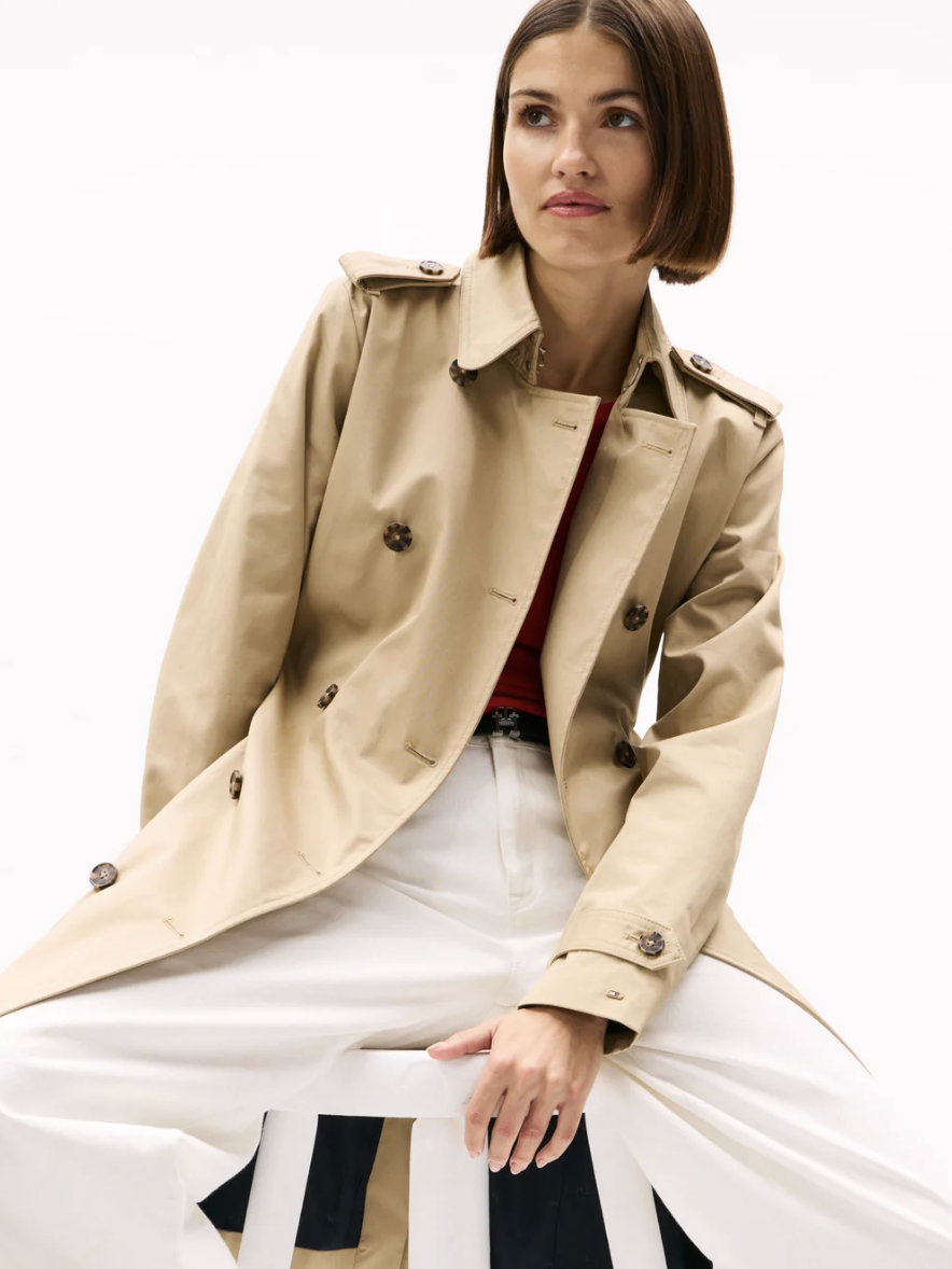 Woman with chin-length brown hair sitting on a white stool against a plain background, wearing a beige trench coat, white pants, and a red top.