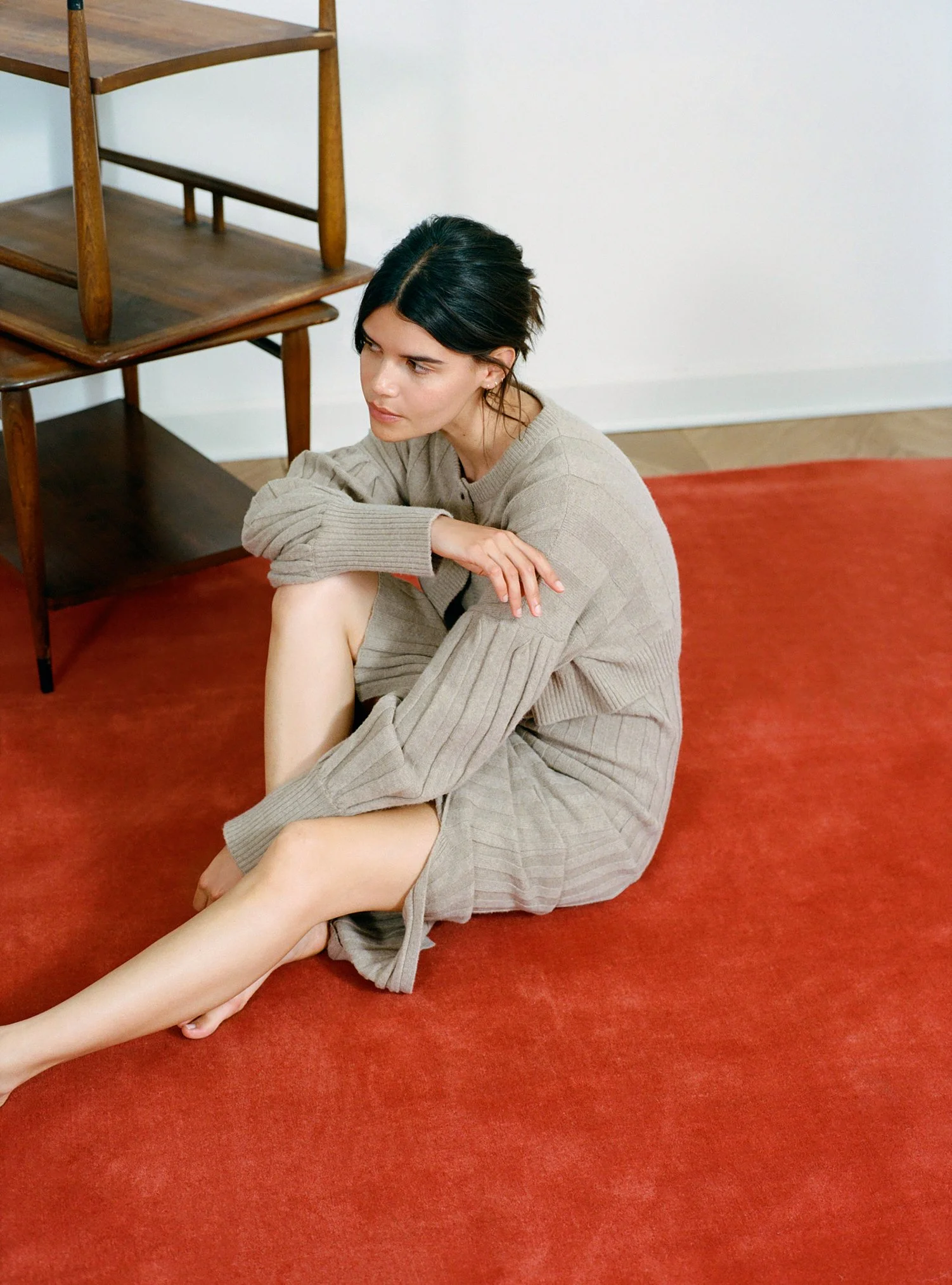 A woman with black hair sits on a red carpeted floor, wearing a beige sweater and dress, with a contemplative expression, next to a wooden chair and furniture in a minimalist room.