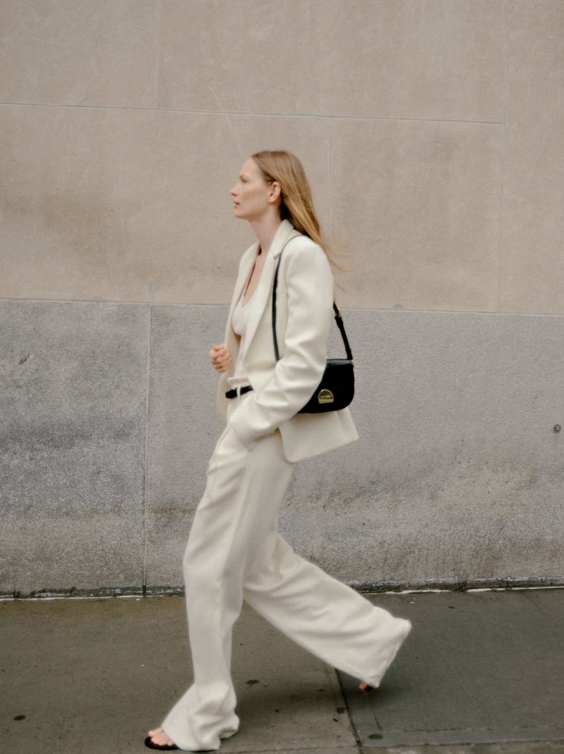 A woman with long red hair wearing a white suit and black sandals, walking along a city sidewalk with a beige stone wall in the background, carrying a black shoulder bag.