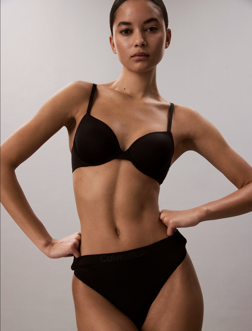 A woman with a confident pose wearing a black bra and matching Calvin Klein underwear, standing against a plain background.