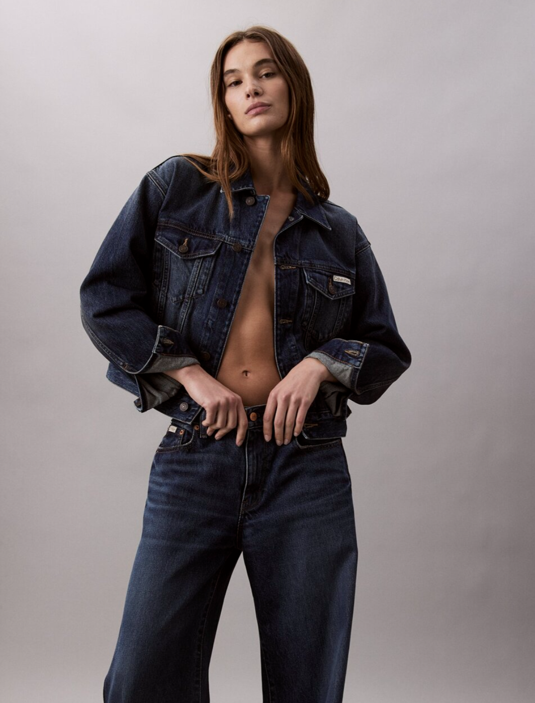 A woman with long brown hair wearing a dark denim jacket and jeans, with the jacket open to reveal her midsection, standing against a plain light textured background.
