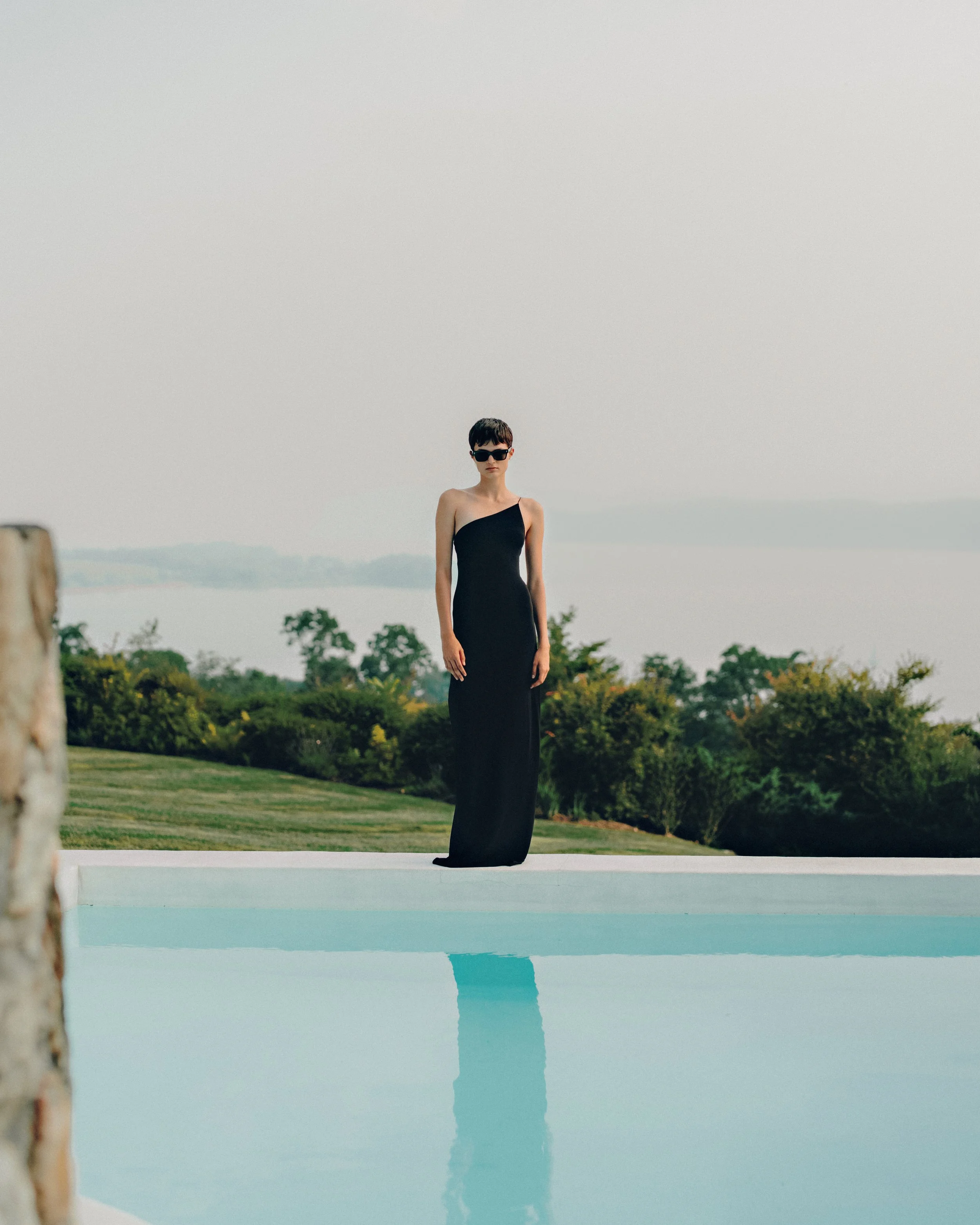 Woman in a long black dress and sunglasses standing near a swimming pool outdoors.