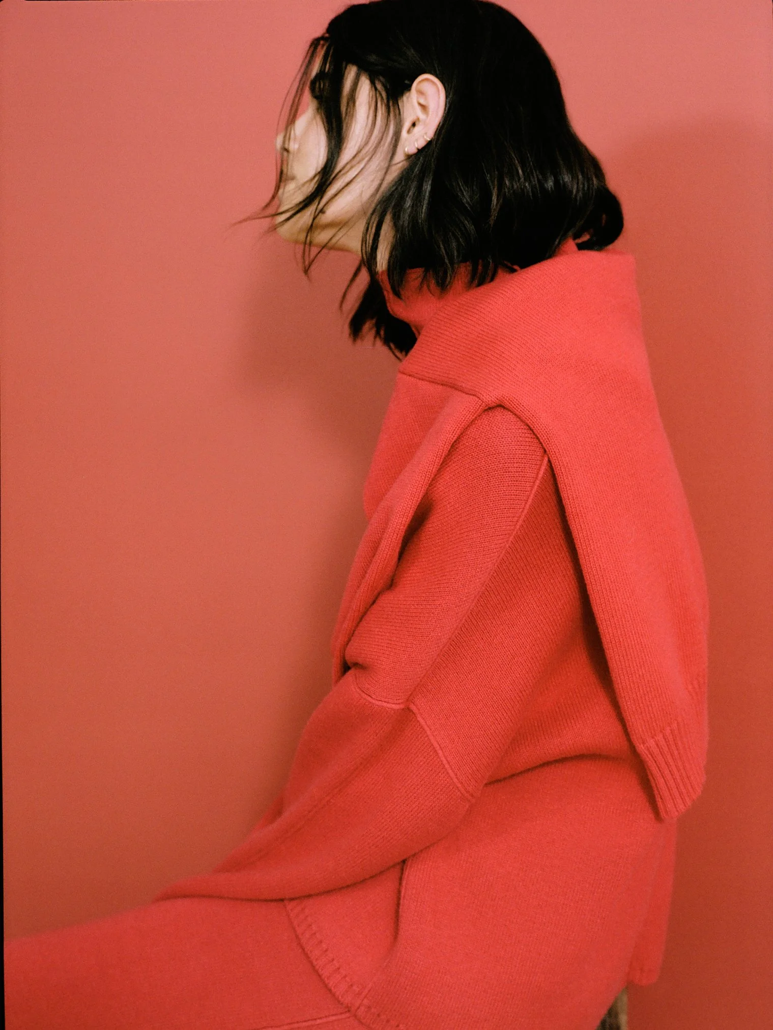 A woman with dark, wavy hair wearing a matching coral-colored turtleneck and oversized sweater, sitting against a coral background with her eyes closed and head slightly tilted.