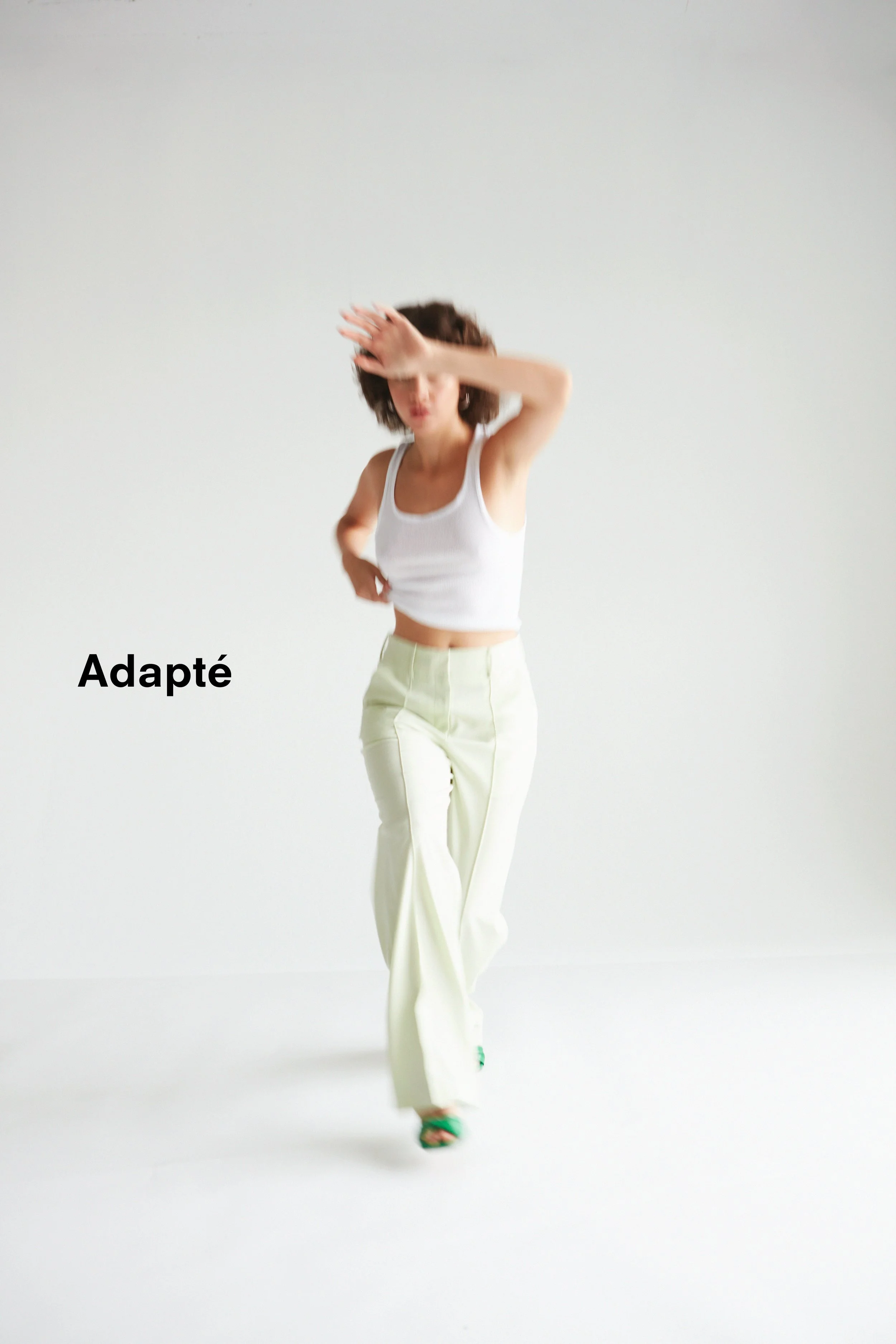 A woman in a white sleeveless crop top and light-colored pants walking forward in a minimalist white space, partially covering her face with her right arm, with the word "Adapté" in bold black text on the left side.