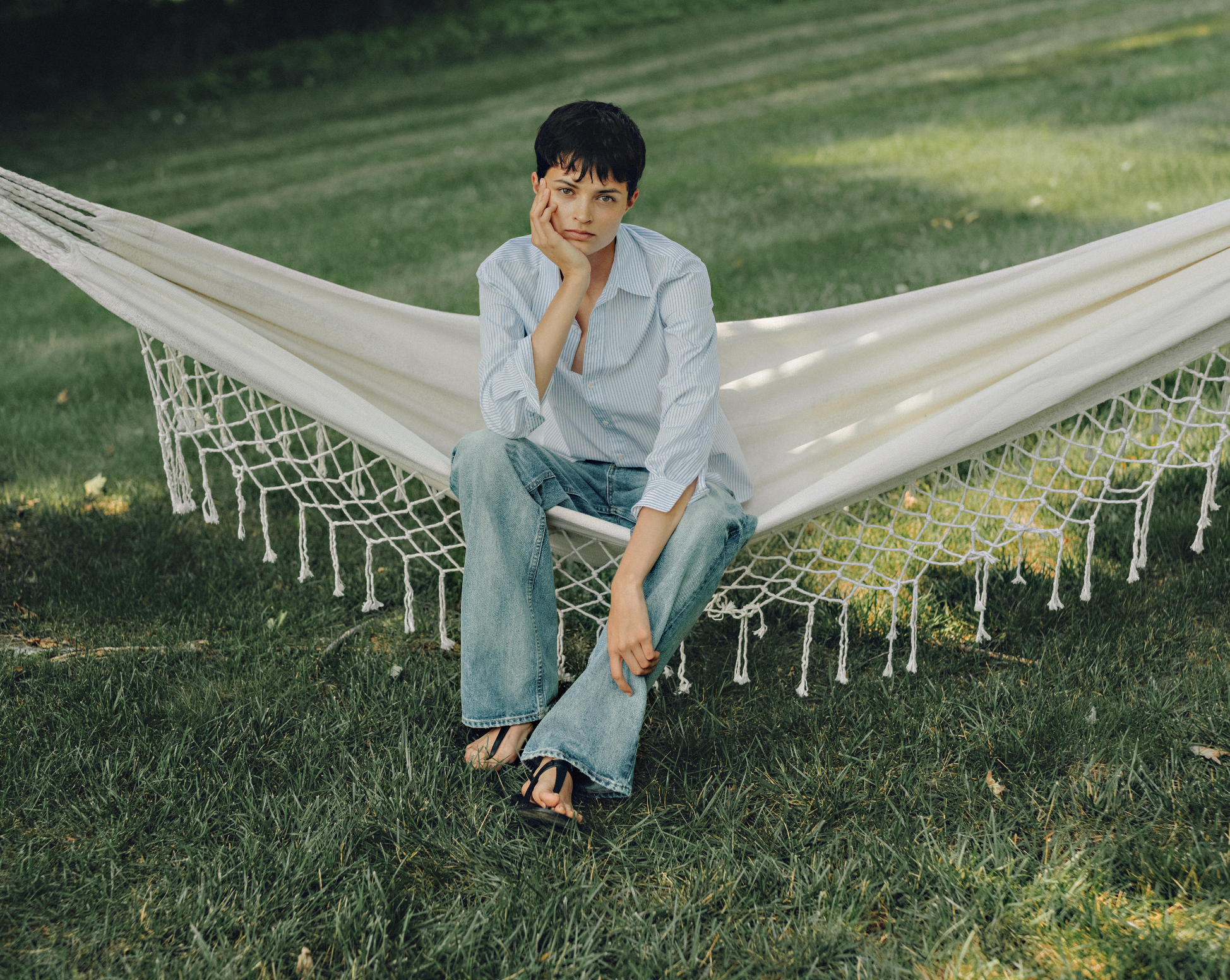 A young woman with short dark hair sitting on a hammock outdoors on a grassy area, wearing a light blue striped shirt, loose jeans, and flip-flops, with a pensive expression, resting her face on her hand.