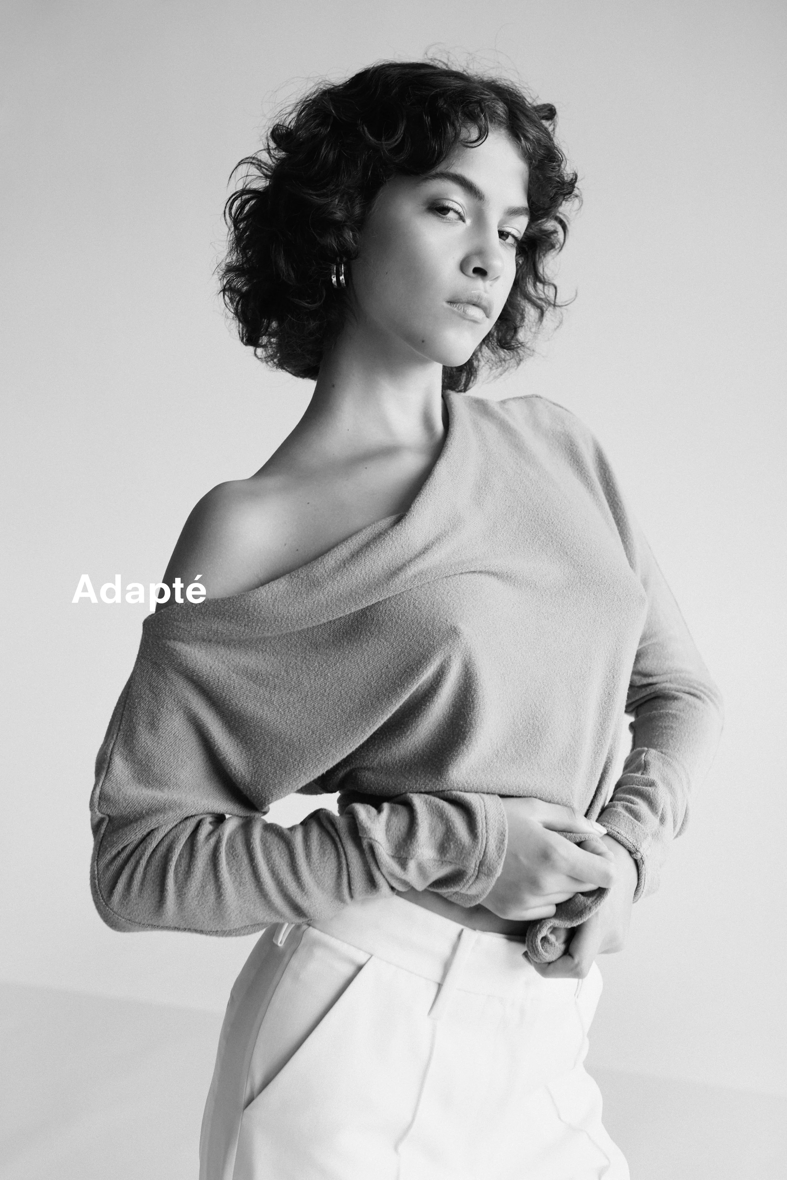 A black and white portrait of a woman with curly hair, wearing a one-shoulder top and high-waisted pants, with the word 'Adapté' overlaid.