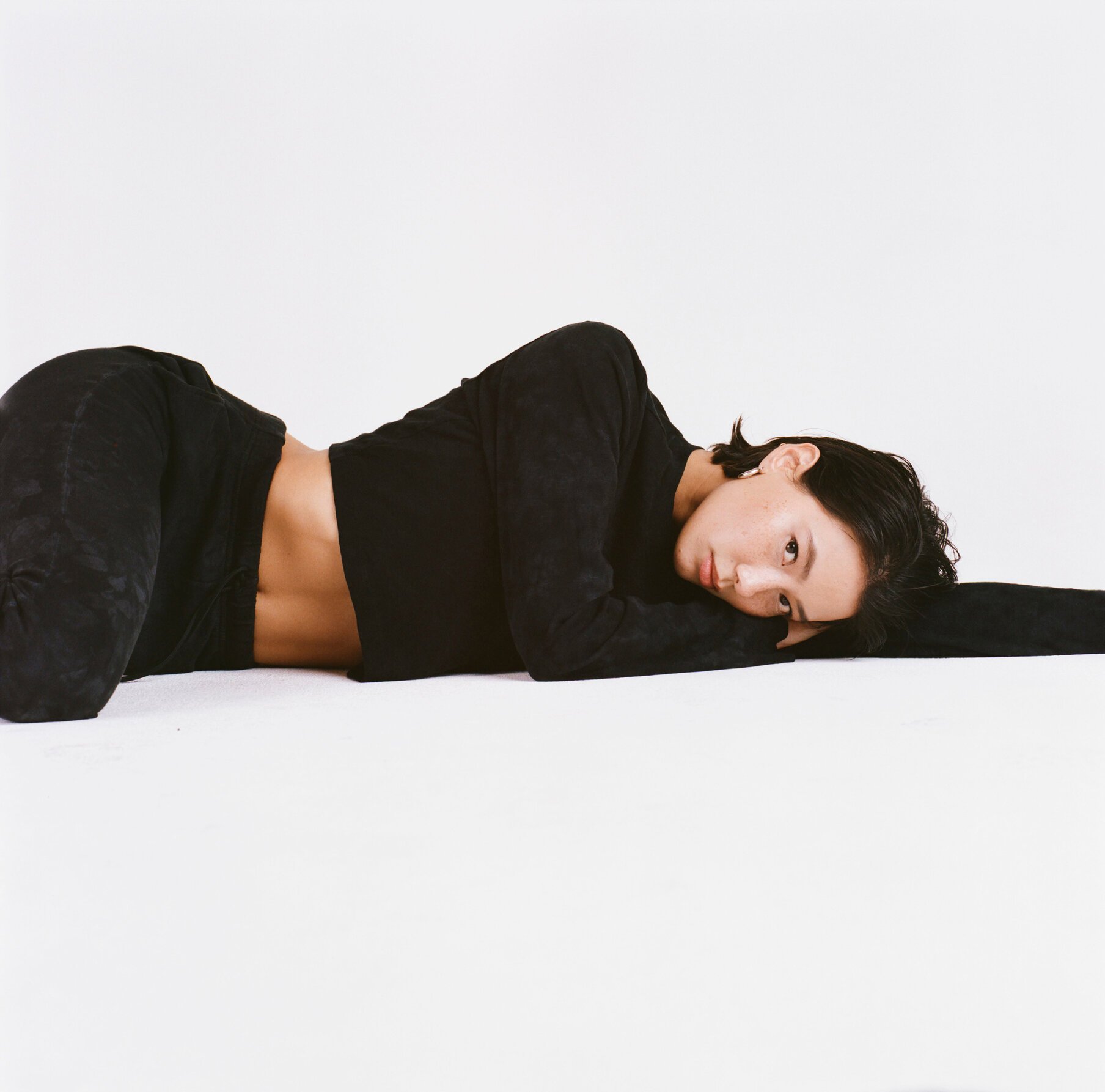 A woman lying on her side against a white background, wearing a black jacket and black pants, resting her head on her arm and gazing at the camera.