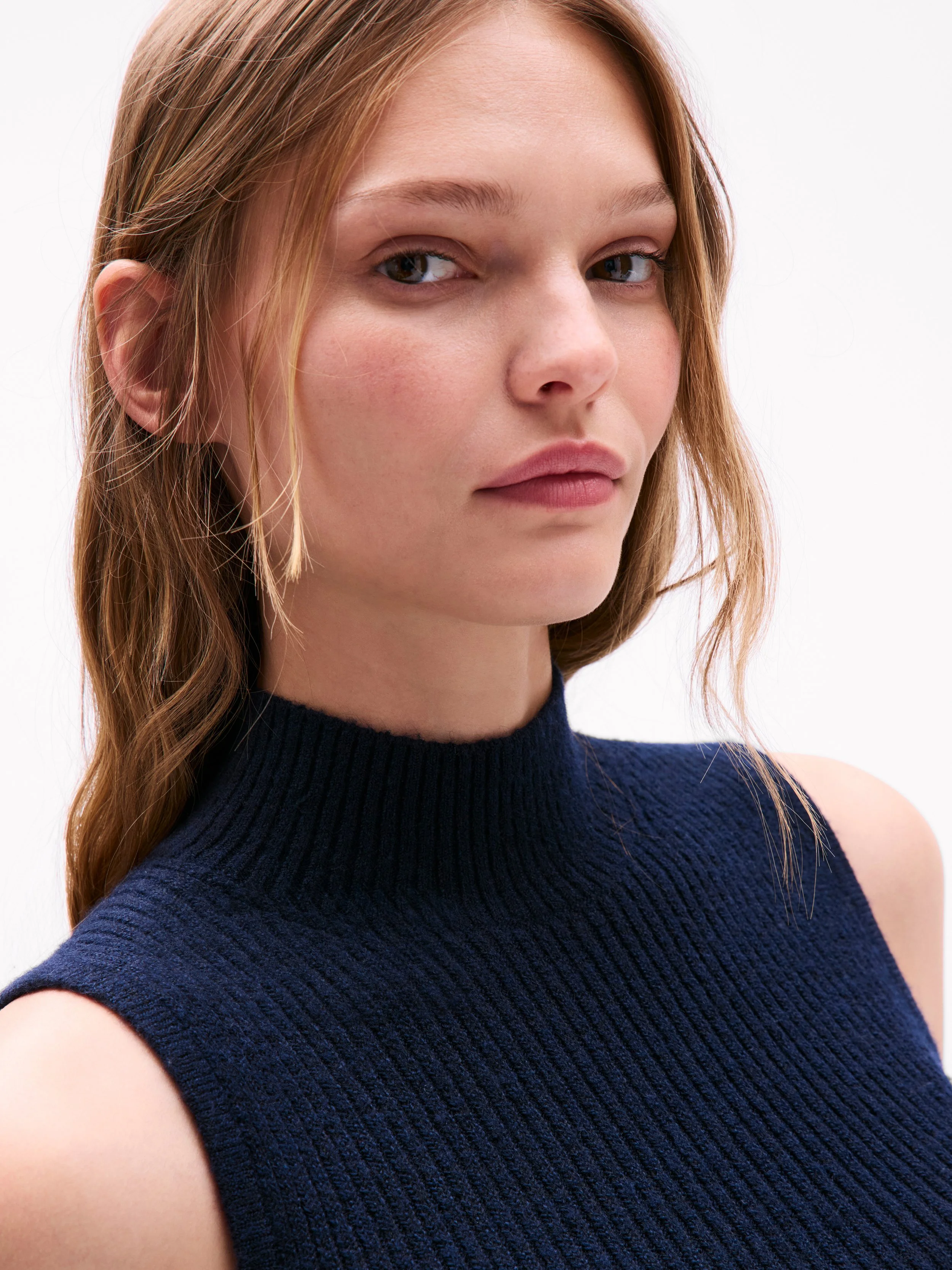 A young woman with light skin and light brown hair with wavy strands, wearing a sleeveless dark blue knit turtleneck sweater, looking at the camera with a neutral expression against a plain white background.