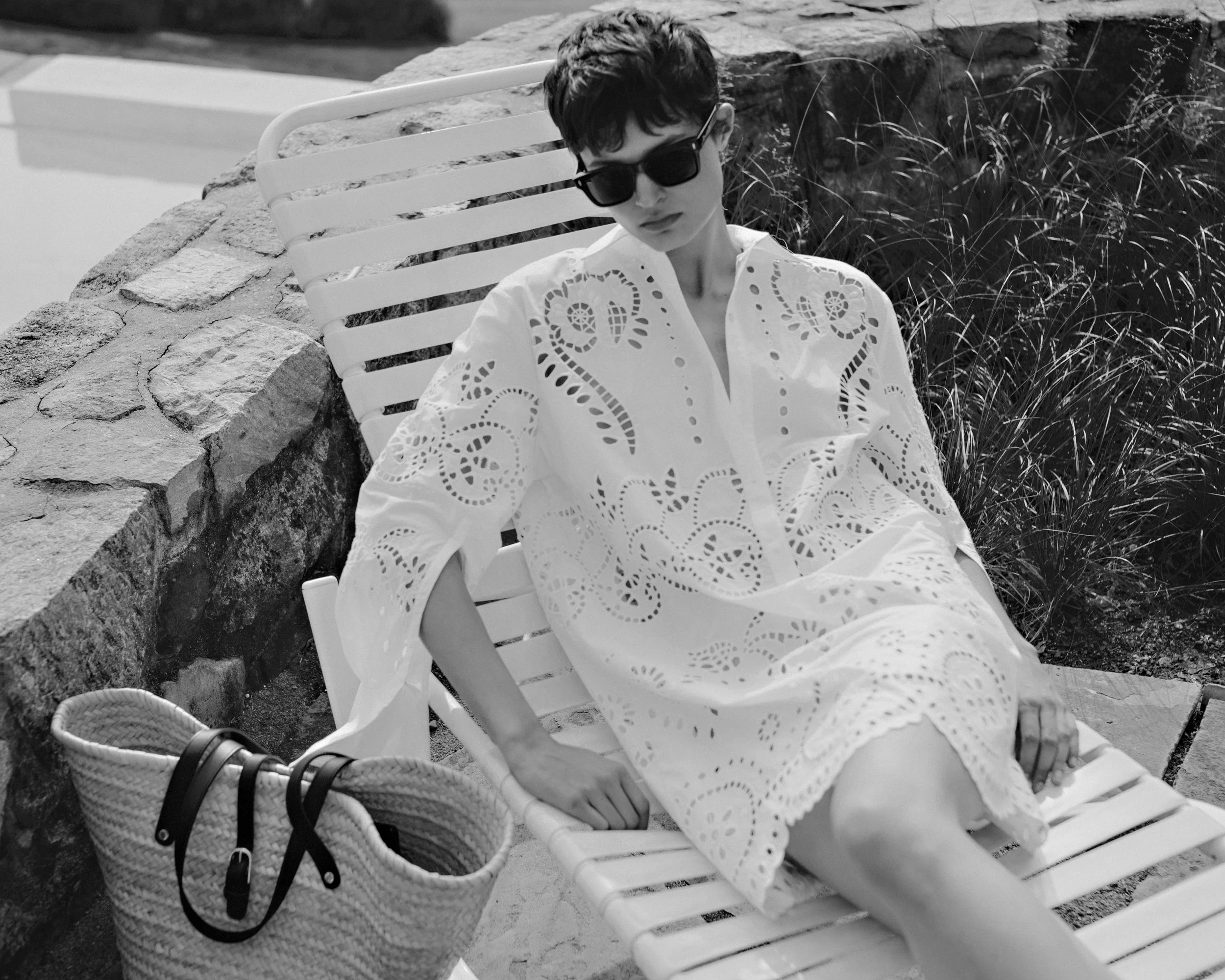 A woman with short hair and sunglasses reclining on a lounge chair outdoors, wearing a white eyelet dress and having a bag placed nearby.