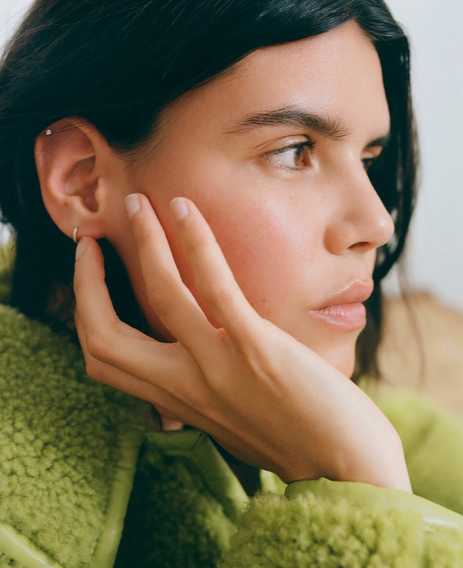 Close-up of a woman with black hair and hoop earrings, resting her face on her hand, wearing a green textured coat.