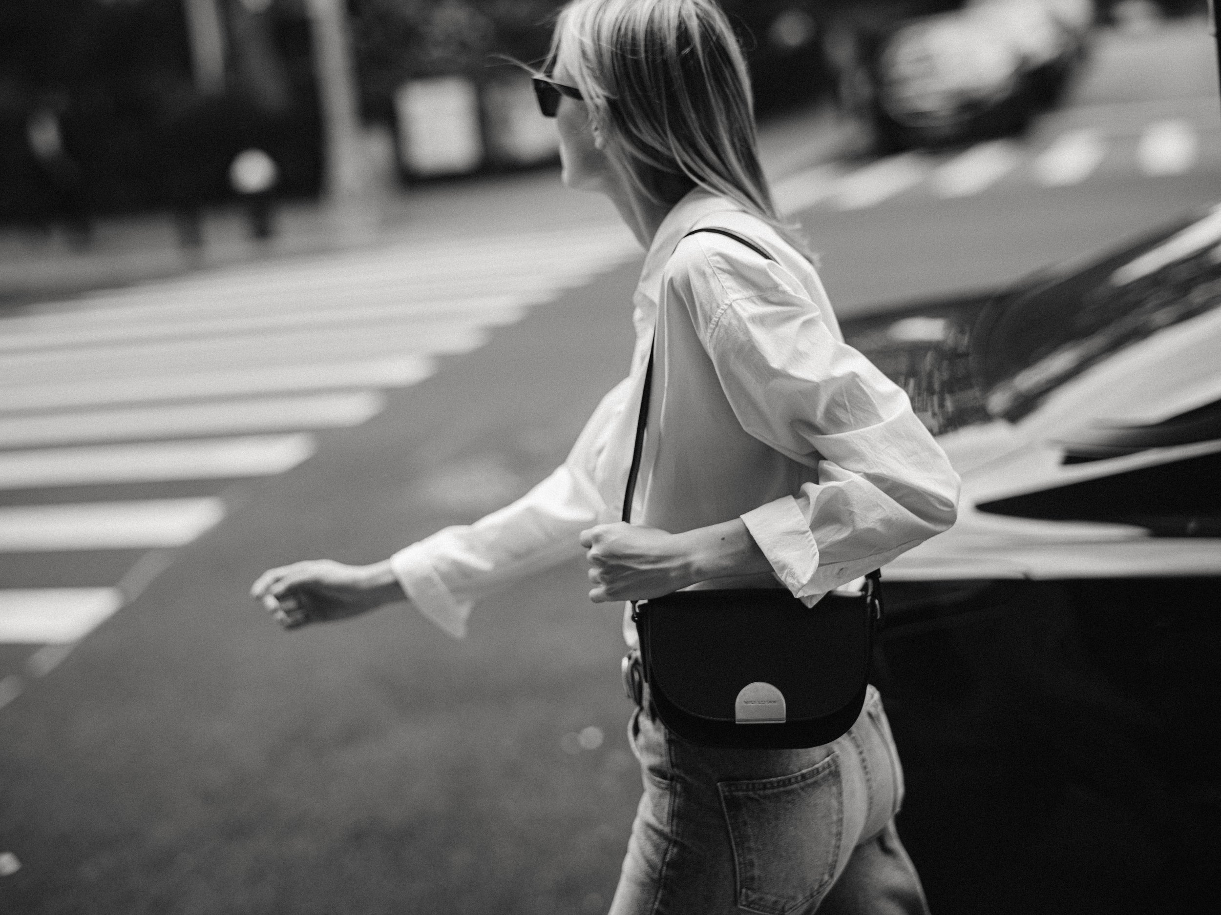 A woman crossing a street at a crosswalk, wearing a white shirt, jeans, sunglasses, and carrying a small shoulder bag.