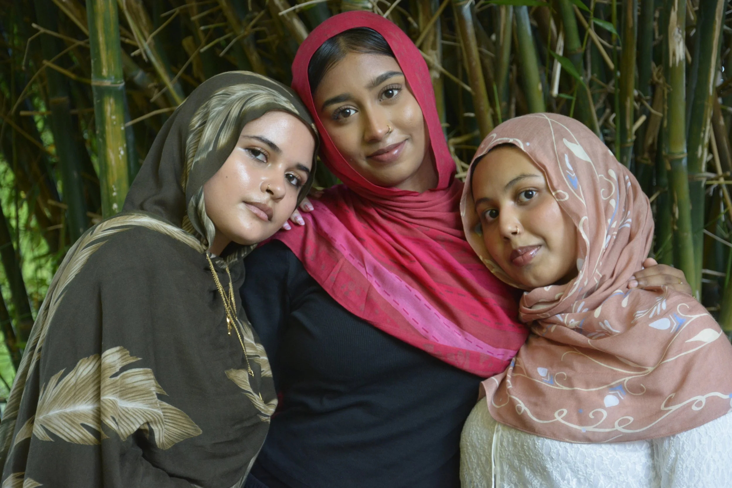 Three women of South Asian descent in colorful traditional attire, smiling and embracing each other in front of bamboo plants.