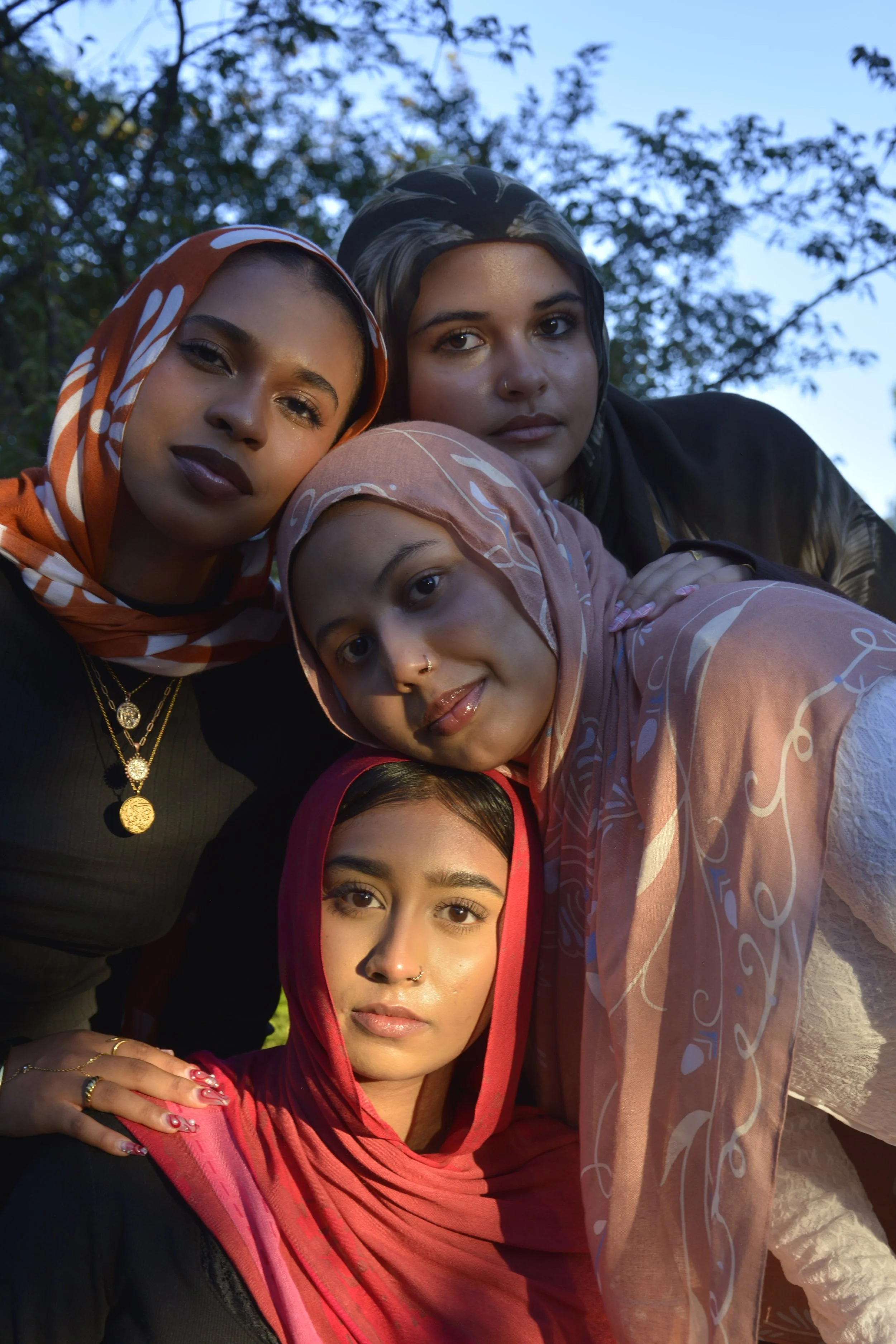 Four women wearing headscarves and jewelry posing closely outdoors during daytime, with trees and sky in the background.