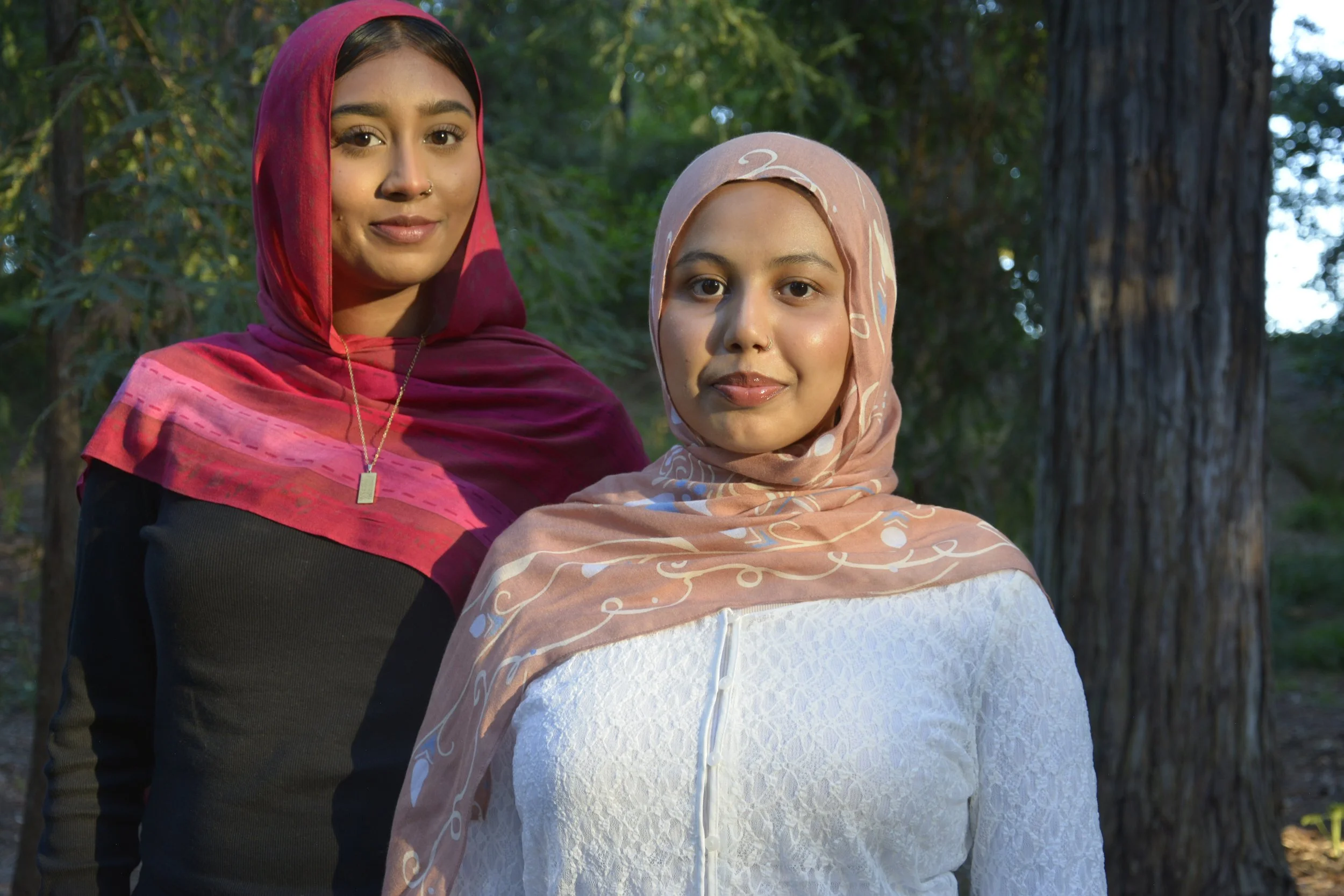 Two women standing outdoors in front of trees, looking at the camera. The woman on the left has light brown skin, wearing a pink headscarf, and the woman on the right has light brown skin, wearing a beige headscarf with white patterns, and a white top.