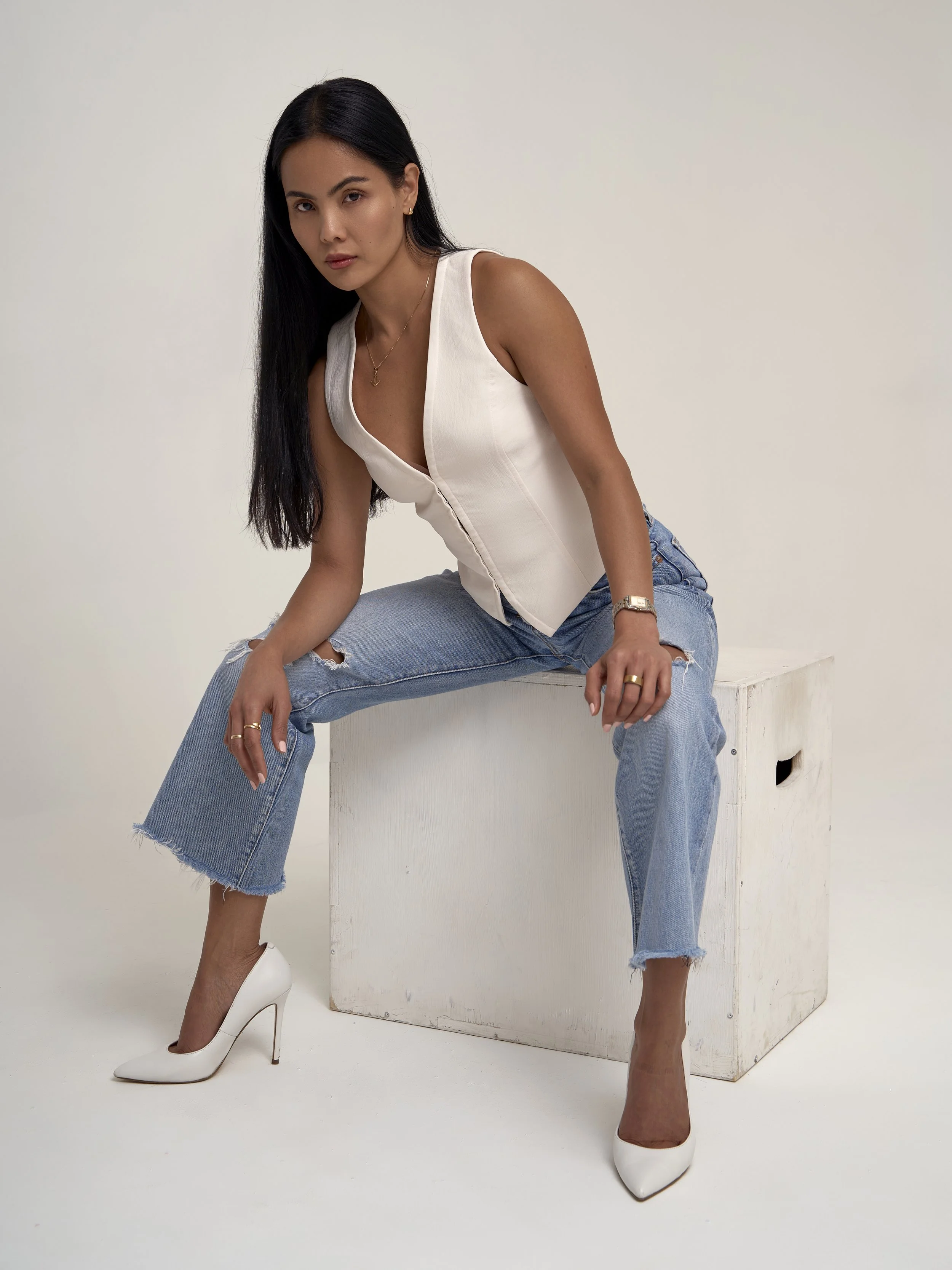 A woman with long black hair, wearing a sleeveless white top, ripped blue jeans, and white high heels, sitting on a white box against a plain background.