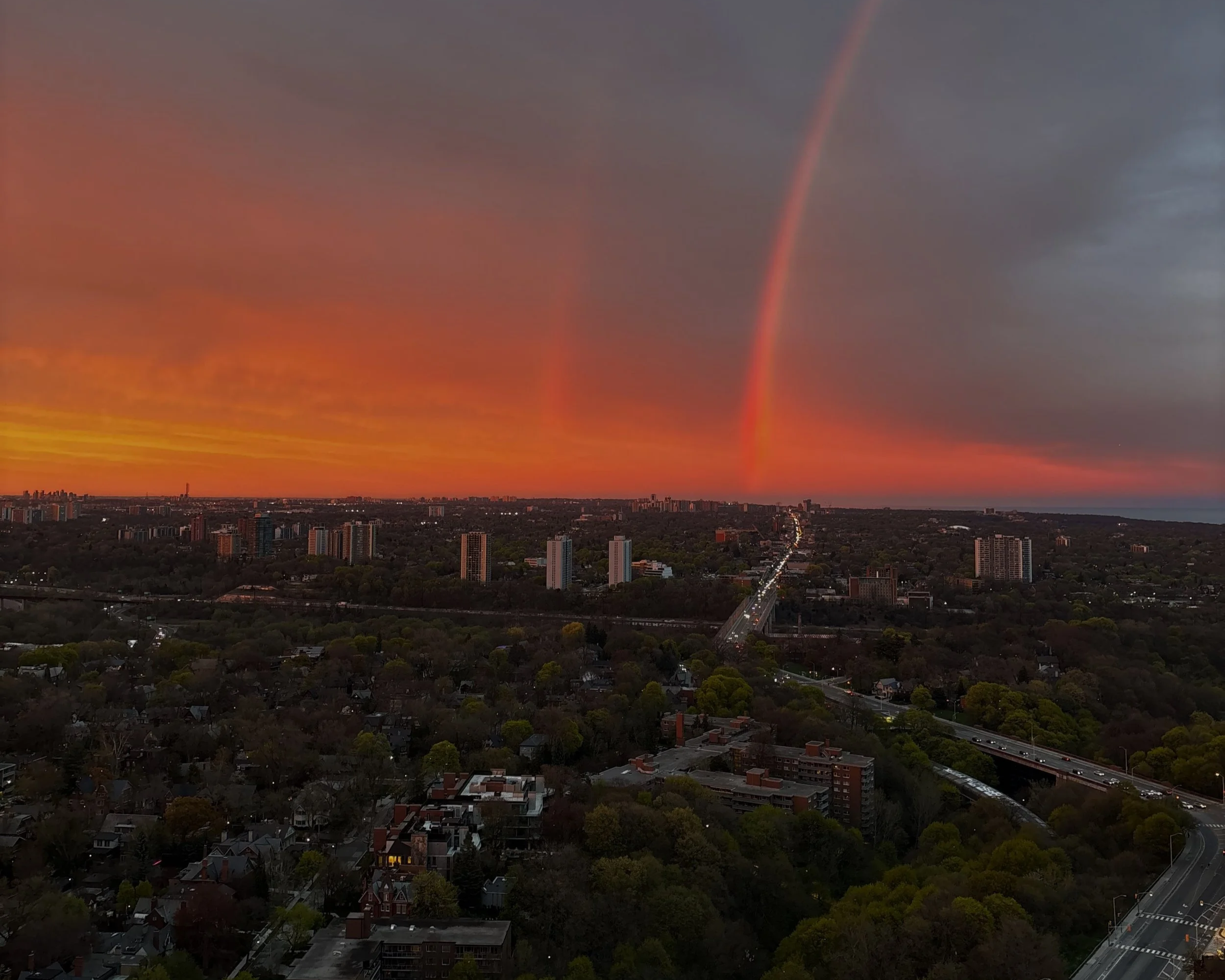Cityscape during sunset with dark clouds and two visible rainbows in the sky, a skyline with tall buildings and a tree-lined neighborhood in the foreground.