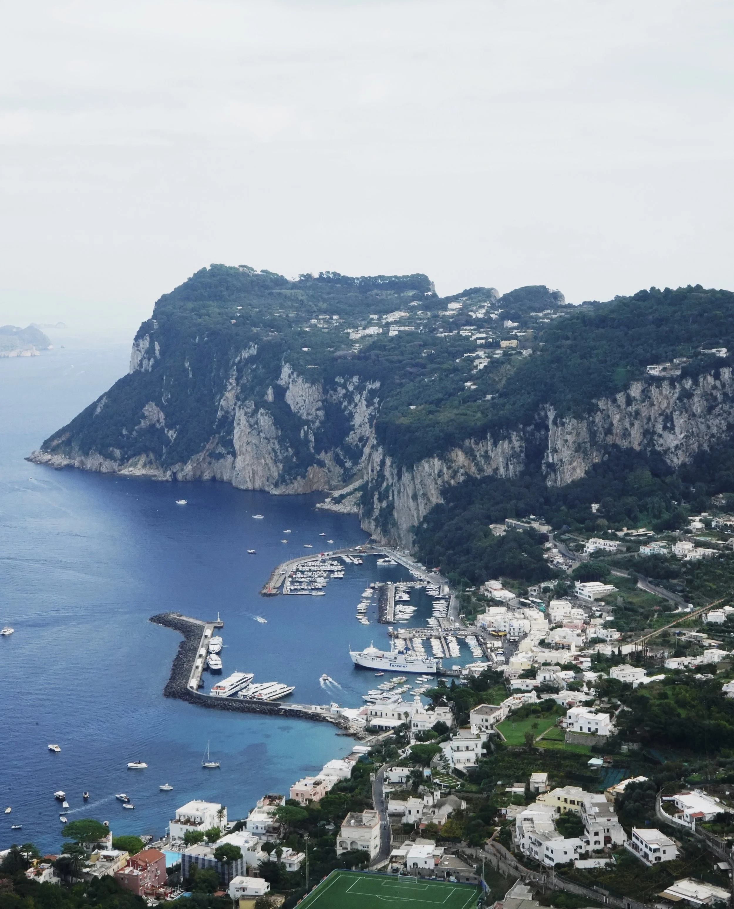 A coastal town with white buildings, a marina with boats, and steep green cliffs in the background.