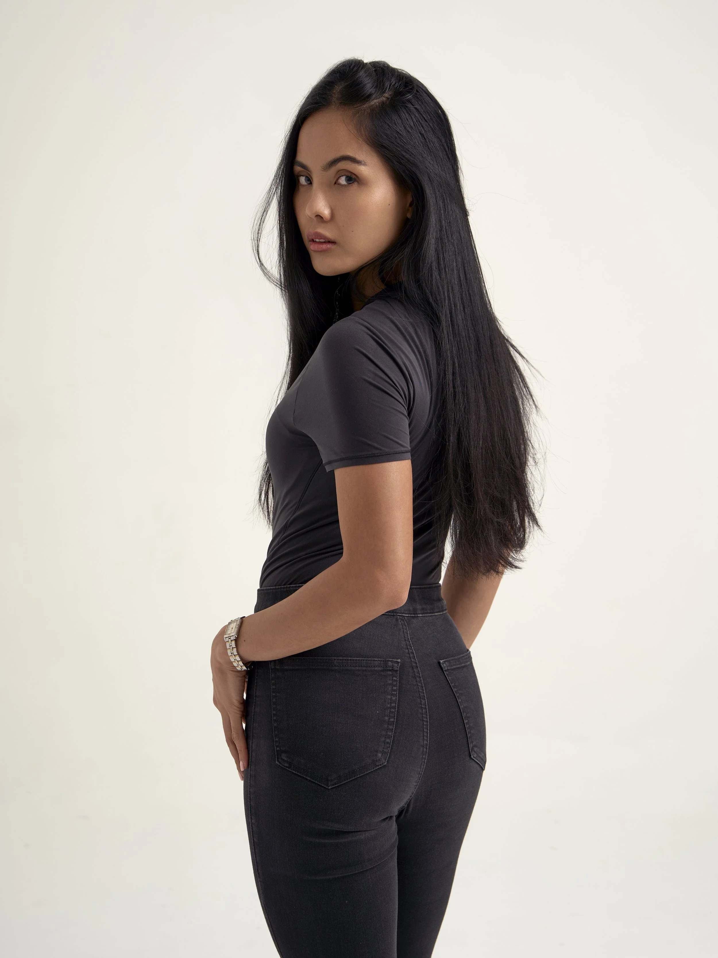 A woman with long black hair wearing a fitted black shirt and black jeans, standing against a plain light-colored background.