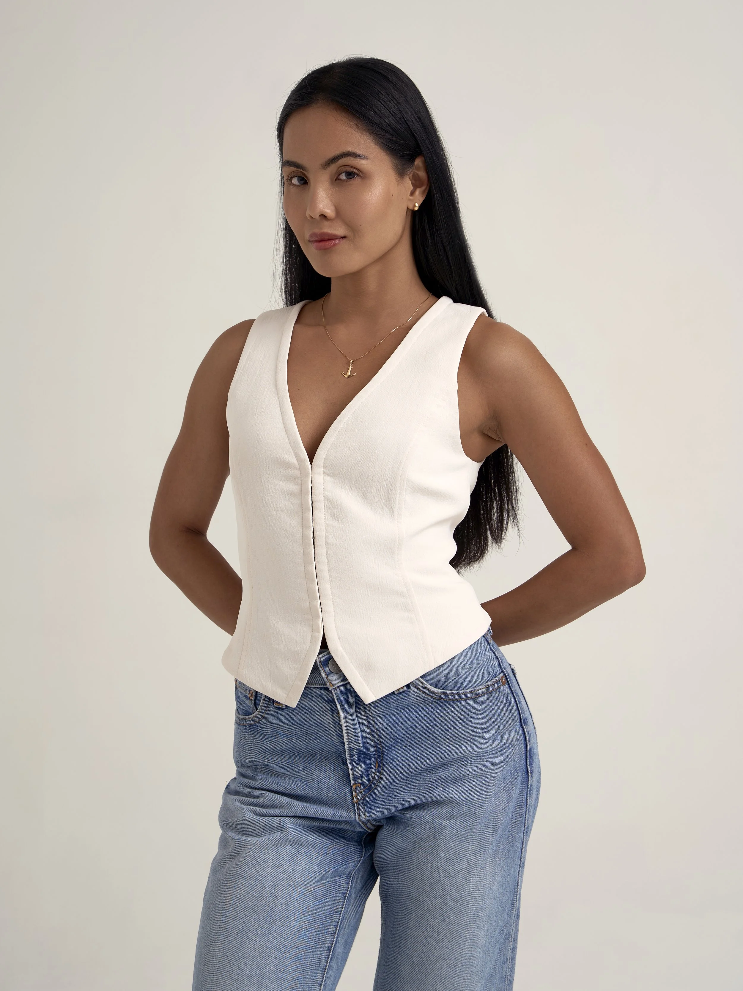 A woman with long, dark hair posing against a plain white background. She is wearing a sleeveless white top with a V-neck and light blue jeans, accessorized with a gold necklace and small earrings.