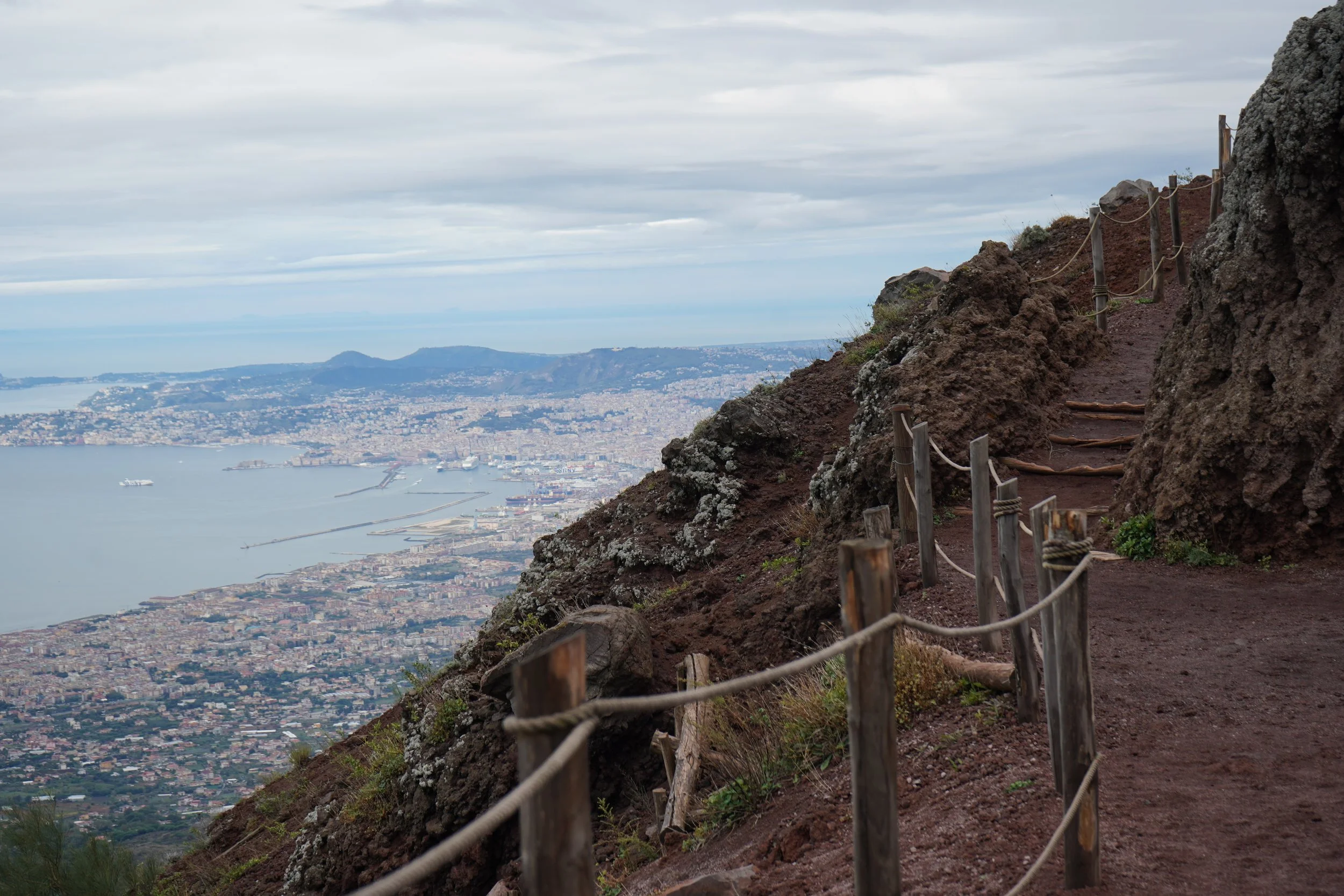 A rocky mountain trail with a wooden rope barrier, overlooking a coastal cityscape with ships, a bay, and distant mountains under a cloudy sky.