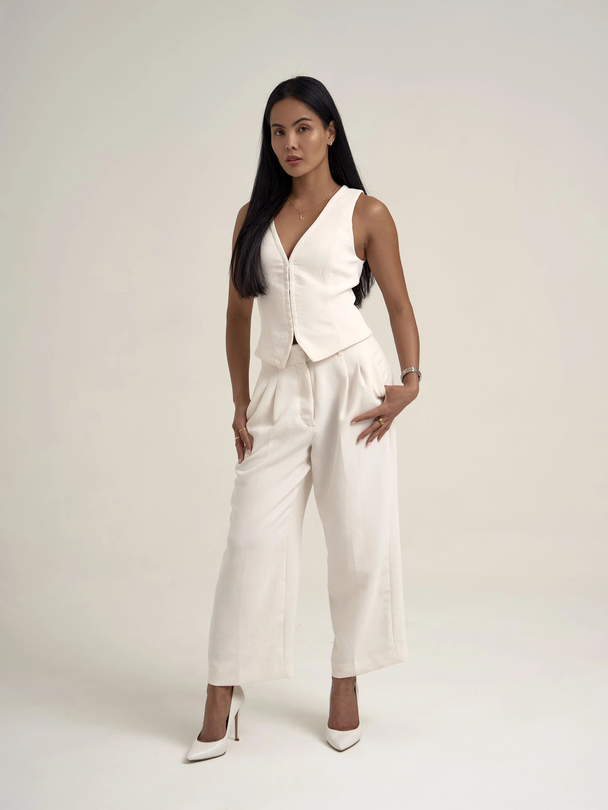 Woman in cream-colored sleeveless vest and wide-leg pants, wearing white high heels, standing against a plain background.