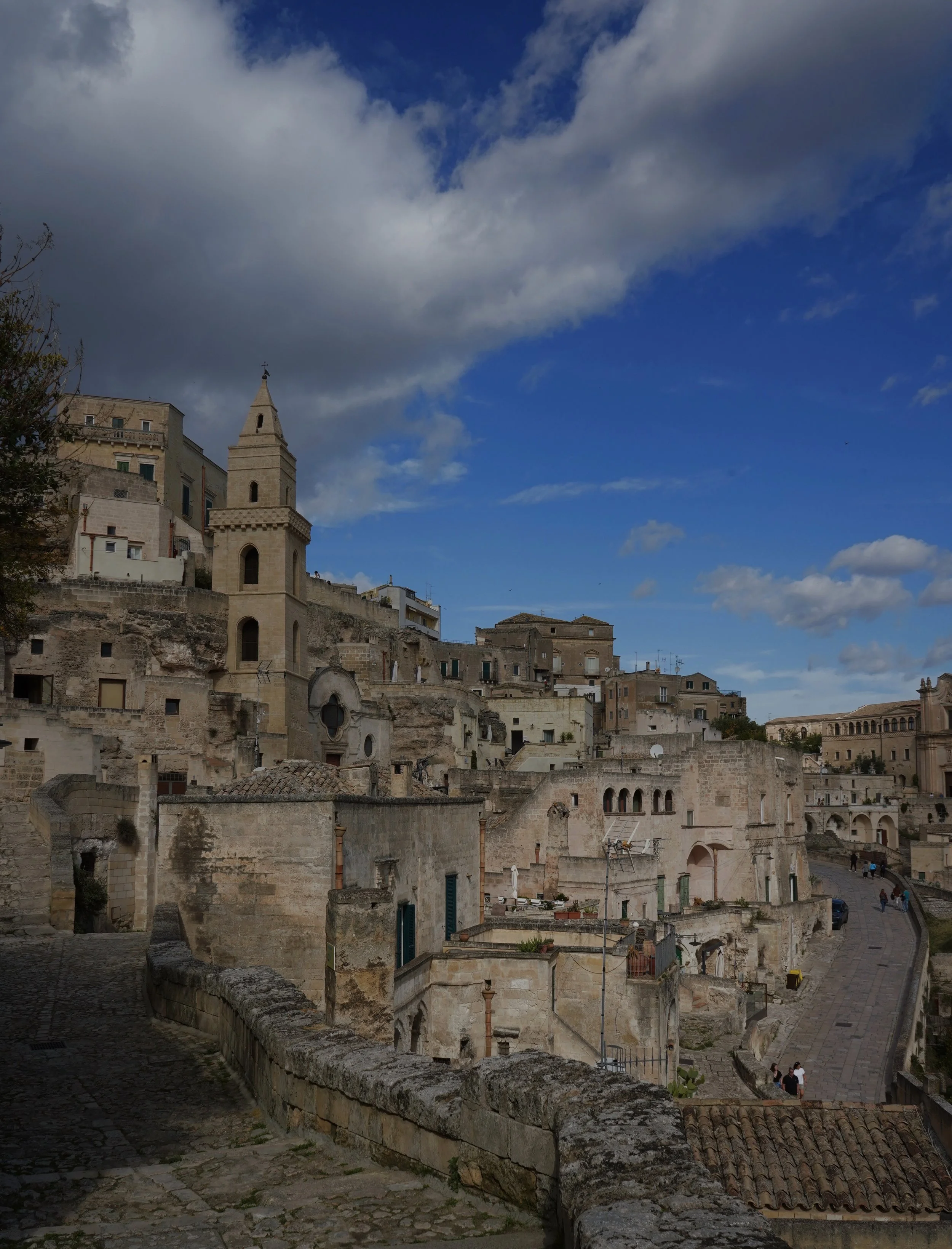 View of ancient stone buildings and a church with a bell tower in a hillside town under a partly cloudy sky.