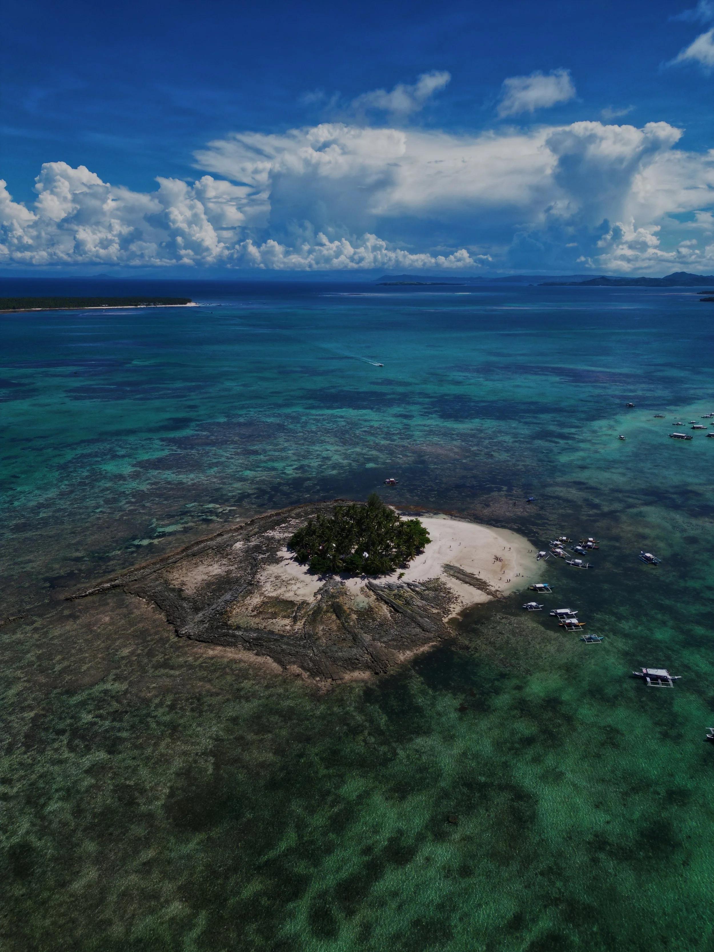 Aerial view of a tropical island with a small beach and green trees, surrounded by turquoise waters and boats, under a partly cloudy sky.
