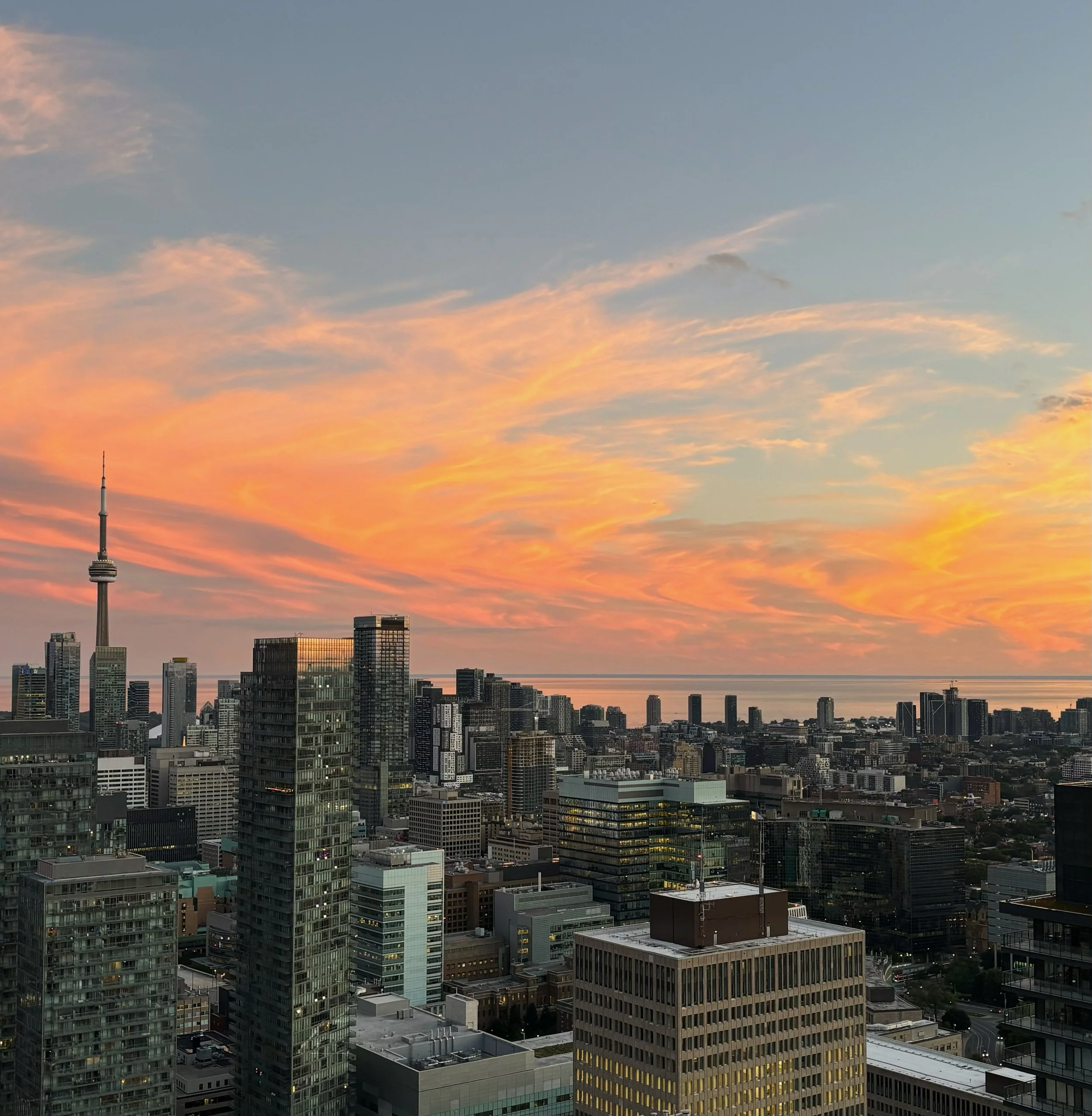 City skyline at sunset with tall buildings and the CN Tower in the distance, under a colorful sky with orange and pink clouds.