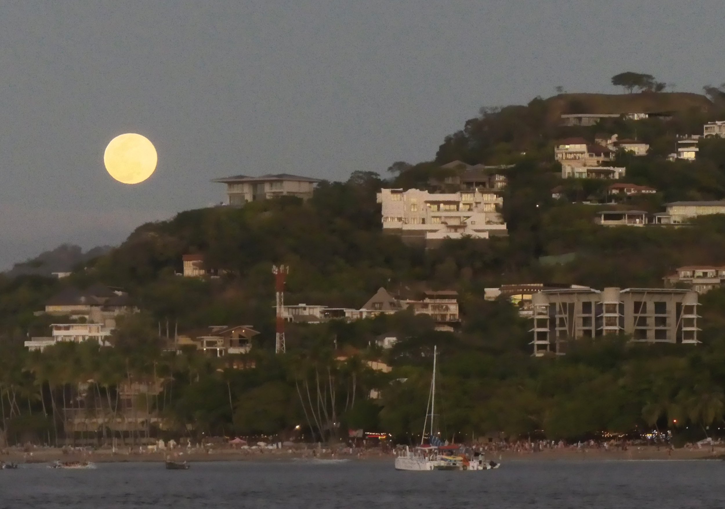 Nighttime view of a hillside with houses and buildings, a full moon in the sky, and boats on the water in the foreground.