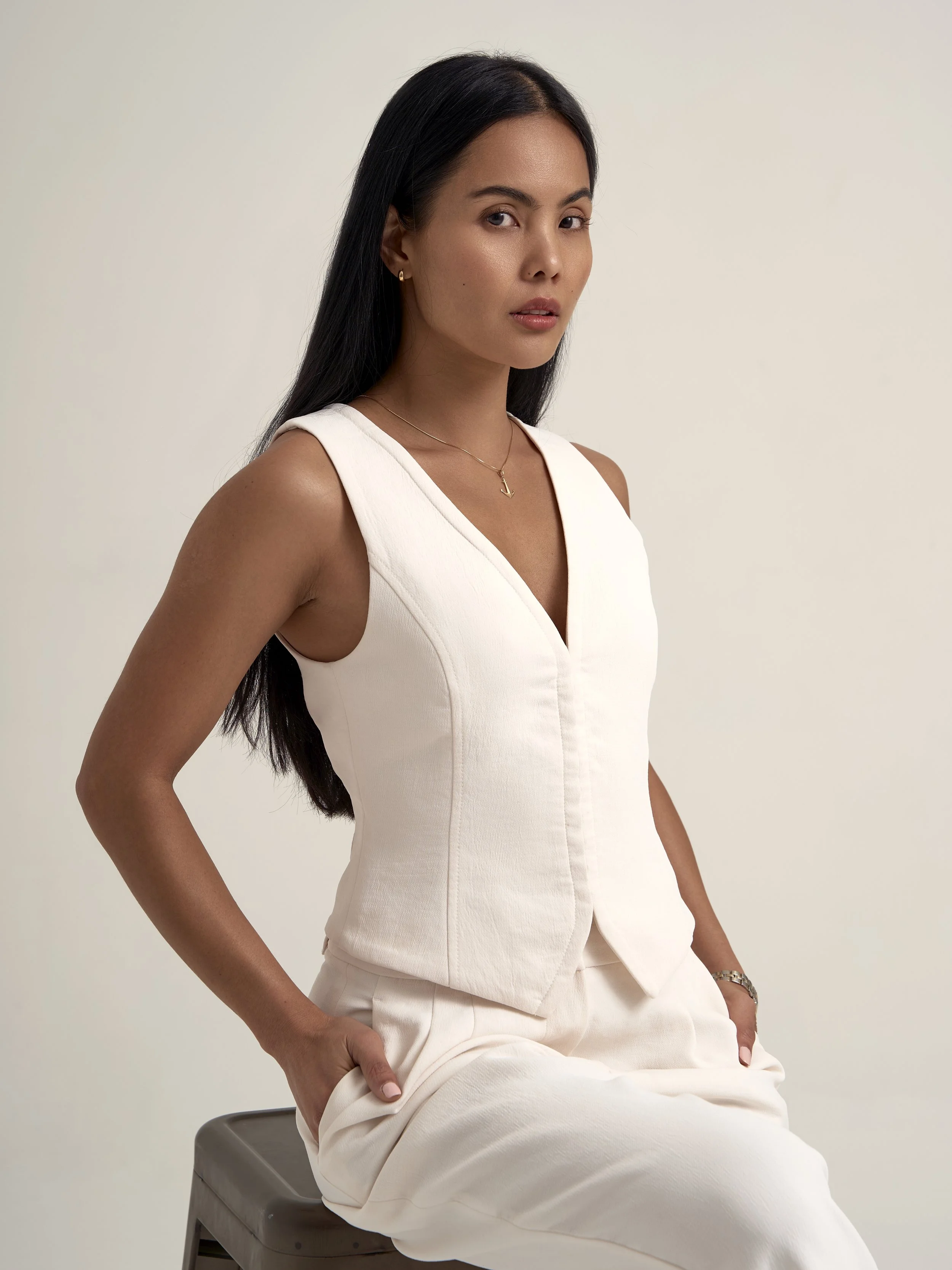 A woman wearing a white sleeveless top and white pants, sitting on a stool with her hands in her pockets, against a plain off-white background.