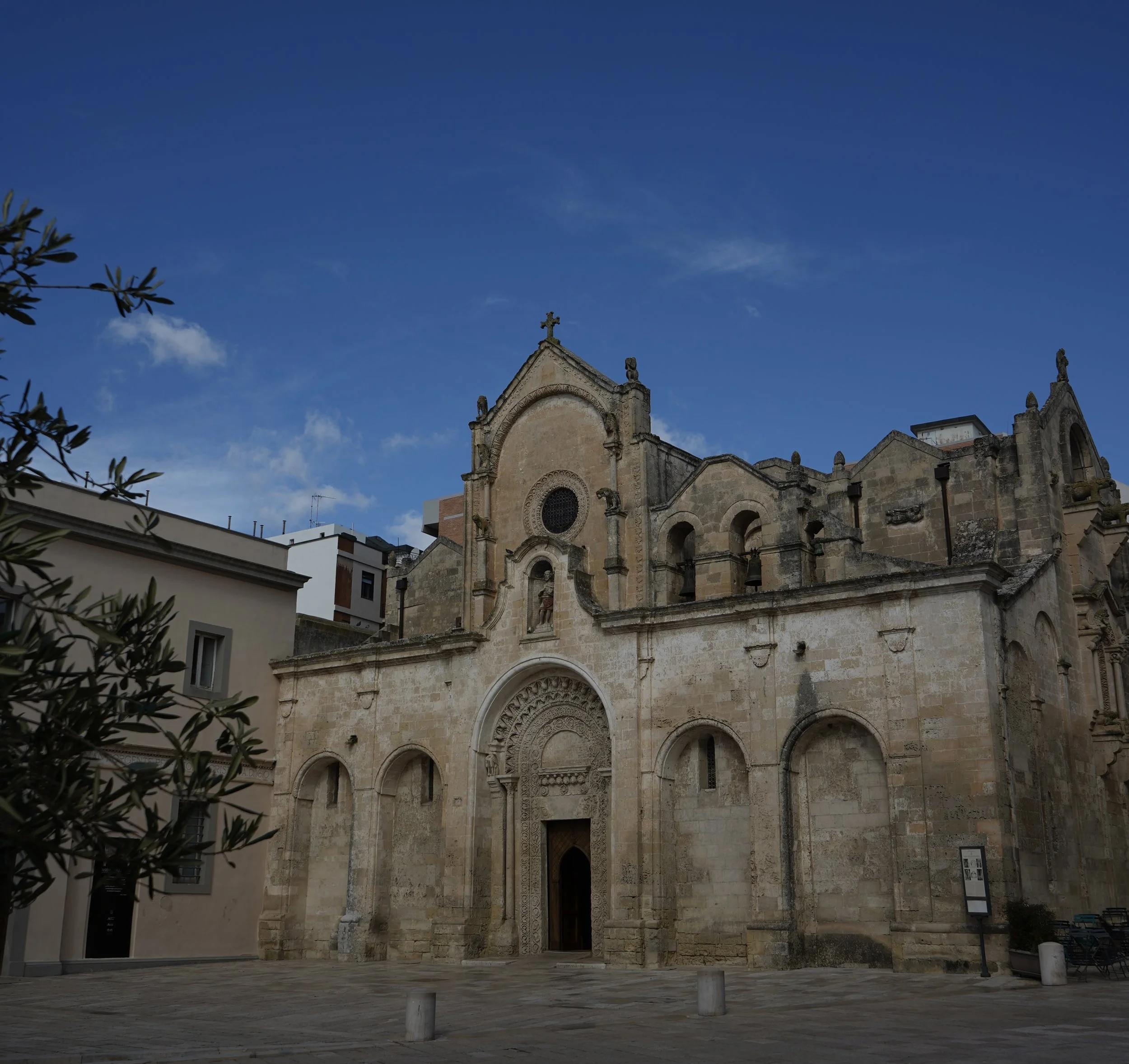 Historic stone church with gothic architectural details, situated in a small square with a few benches and a notice board, under a clear blue sky.